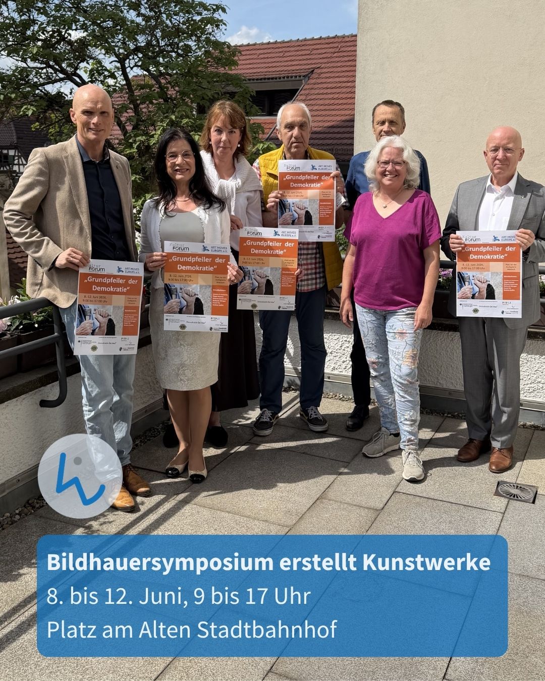 Gruppe von sieben Personen im Freien, die Plakate mit dem Titel „Grundpfeiler der Demokratie“ halten. Im Vordergrund blauer Textblock mit Infos zum Bildhauersymposium vom 8. bis 12. Juni am Platz am Alten Stadtbahnhof. (KI-Alternativtext)