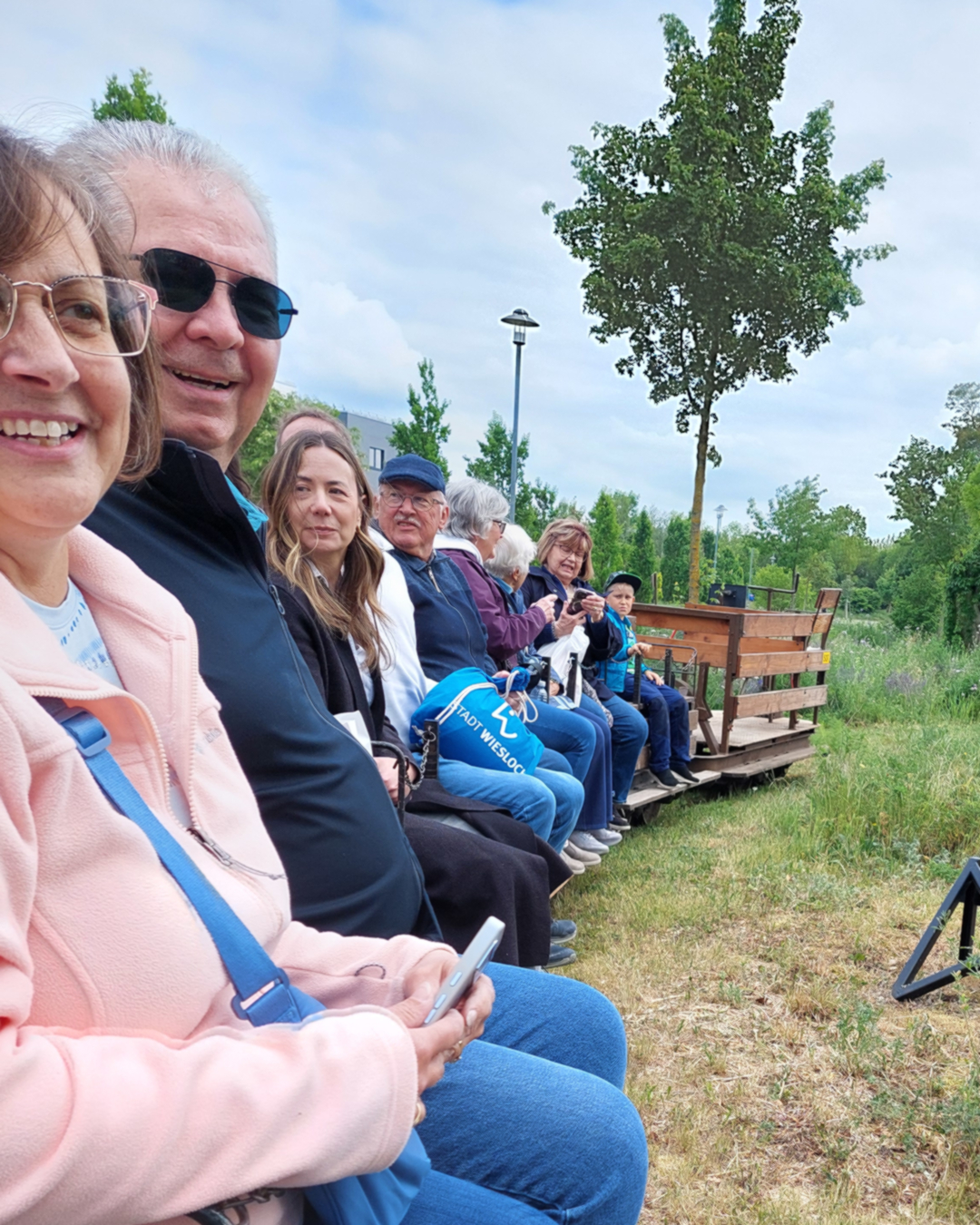 Mehrere Personen sitzen nebeneinander auf einer Holzbank im Freien, einige halten Smartphones, im Hintergrund Bäume und ein blauer Himmel. Eine Person trägt eine blaue Tasche mit der Aufschrift „Stadt Wiesloch“. (KI-Alternativtext)