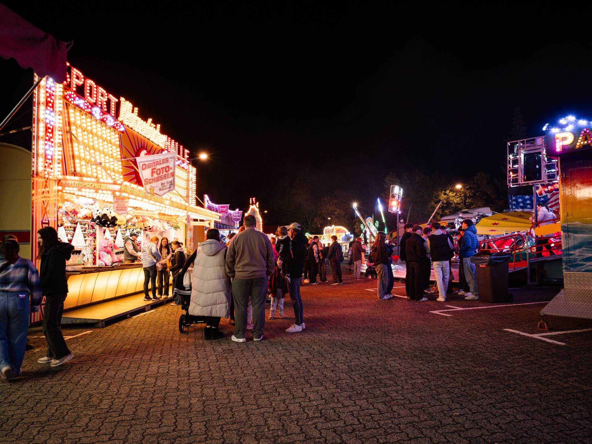 Dieser Alternativtext wurde KI-generiert: Eine belebte Szene bei Nacht, vermutlich auf einem Jahrmarkt oder einer Kirmes. Menschen stehen und bewegen sich zwischen hell erleuchteten Buden und Fahrgeschäften.