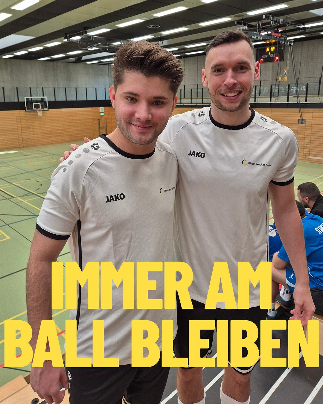 Dieser Alternativtext wurde KI-generiert: Zwei Personen in Sportkleidung stehen lächelnd nebeneinander in einer Sporthalle. Im Hintergrund ist eine Handballhalle erkennbar und ein Banner mit Aufschrift.