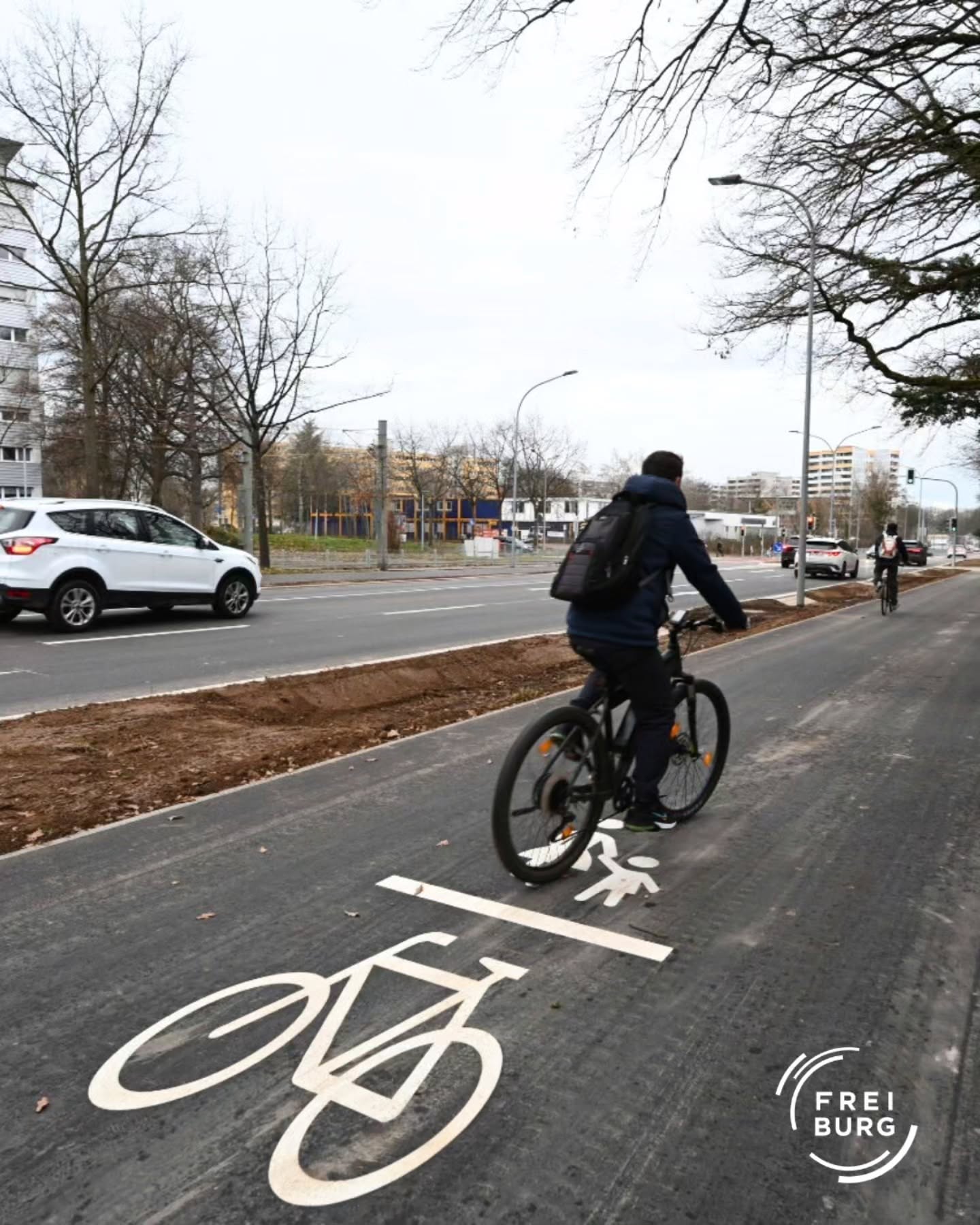 Dieser Alternativtext wurde KI-generiert: Eine Person fährt auf einem Fahrrad auf einem Radweg entlang einer Straße. Im Hintergrund sind Autos und Gebäude zu sehen.