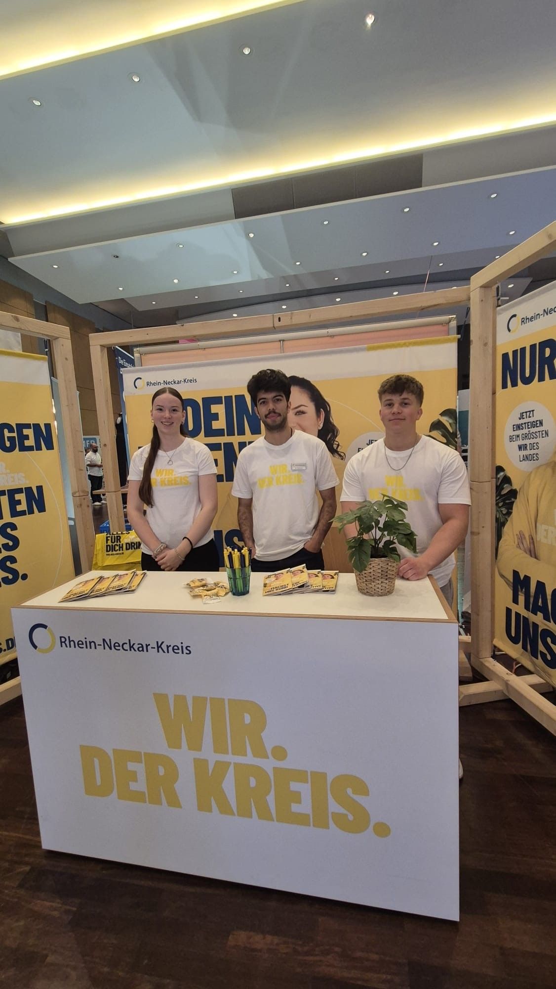 Drei Personen stehen hinter einem weißen Stand mit der Aufschrift "Rhein-Neckar-Kreis" und dem großen gelben Schriftzug "WIR. DER KREIS.". Sie tragen weiße T-Shirts mit gelber Schrift. Auf der Theke liegen Flyer, ein Becher mit Stiften und eine kleine Pflanze, im Hintergrund ist ein gelbes Banner mit dem gleichen Logo zu sehen. (KI-Alternativtext)