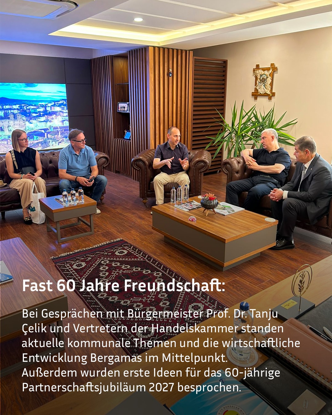 Fünf Personen sitzen in einem modernen Raum auf Ledermöbeln und führen ein Gespräch, wobei eine Person in der Mitte gestikuliert. Ein Textfeld im Vordergrund erläutert, dass bei dem Treffen kommunale Themen, die wirtschaftliche Entwicklung Bergamas sowie das 60-jährige Partnerschaftsjubiläum 2027 besprochen wurden. (KI-Alternativtext)