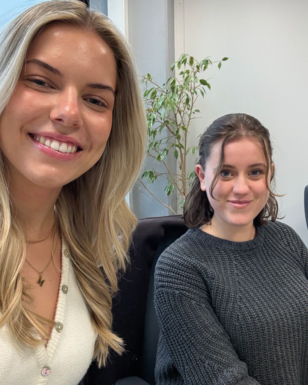 Dieser Alternativtext wurde KI-generiert: Zwei Personen lächeln in die Kamera. Im Hintergrund ist eine Pflanze und ein Bürostuhl zu sehen.