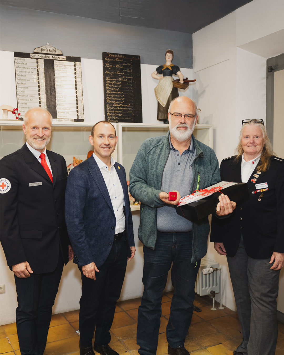 Dieser Alternativtext wurde KI-generiert: Eine Gruppe von vier Personen steht in einem Innenraum vor einer Wand mit Schriften und einer Statue. Eine Person übergibt eine rot-weiße Schachtel an eine andere.