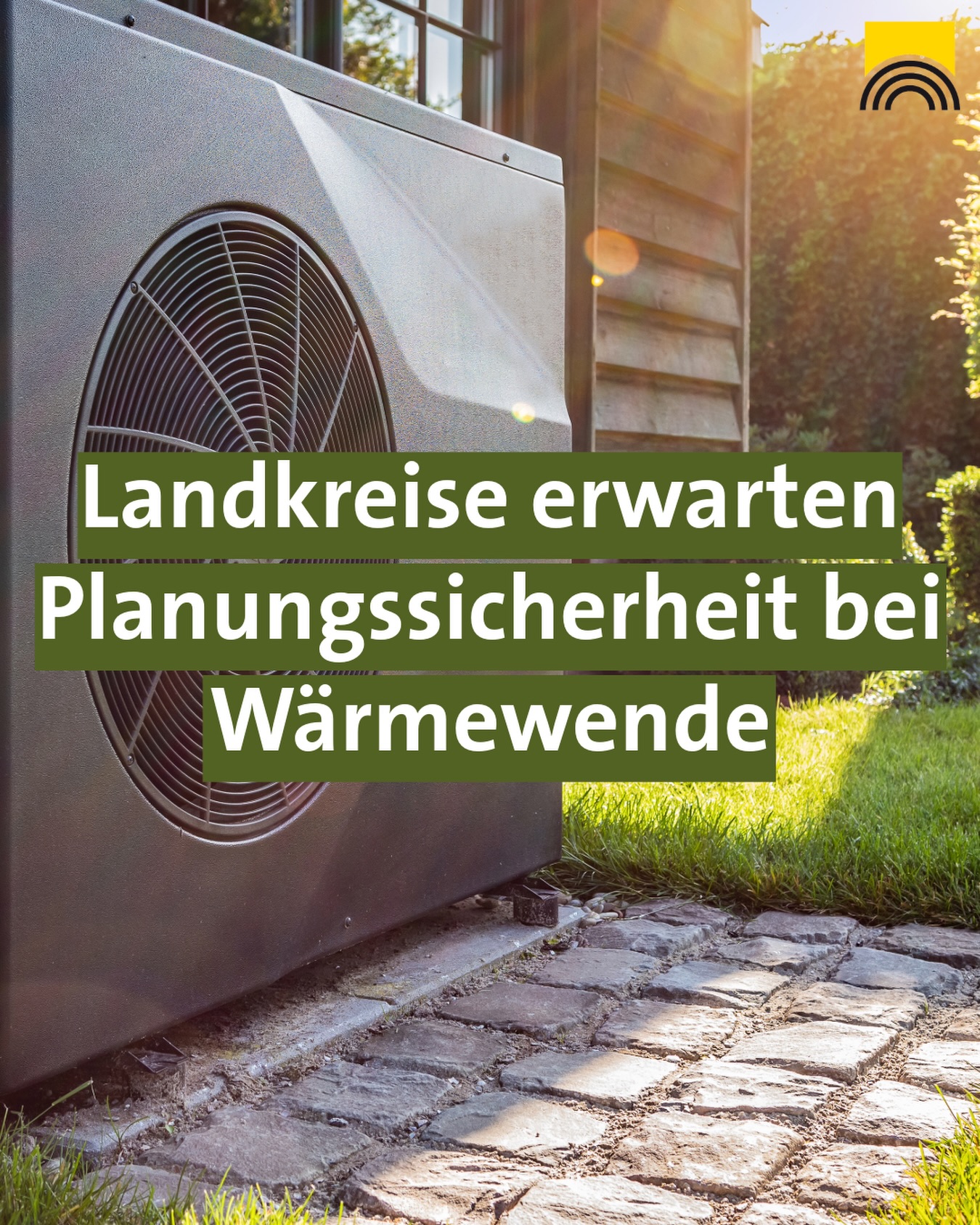 Dieser Alternativtext wurde KI-generiert: Ein großes, graues Gerät mit Lüftungsgitter steht vor einer Hecke und einem Pflasterweg. Über dem Gerät befindet sich ein Textfeld mit Aufschrift.