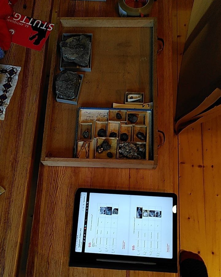 Dieser Alternativtext wurde KI-generiert: Ein Holztisch zeigt eine offene Holzkiste mit vielen kleinen, dunklen Objekten darin. Ein Tablet mit kleinen Bildern liegt vor der Kiste.