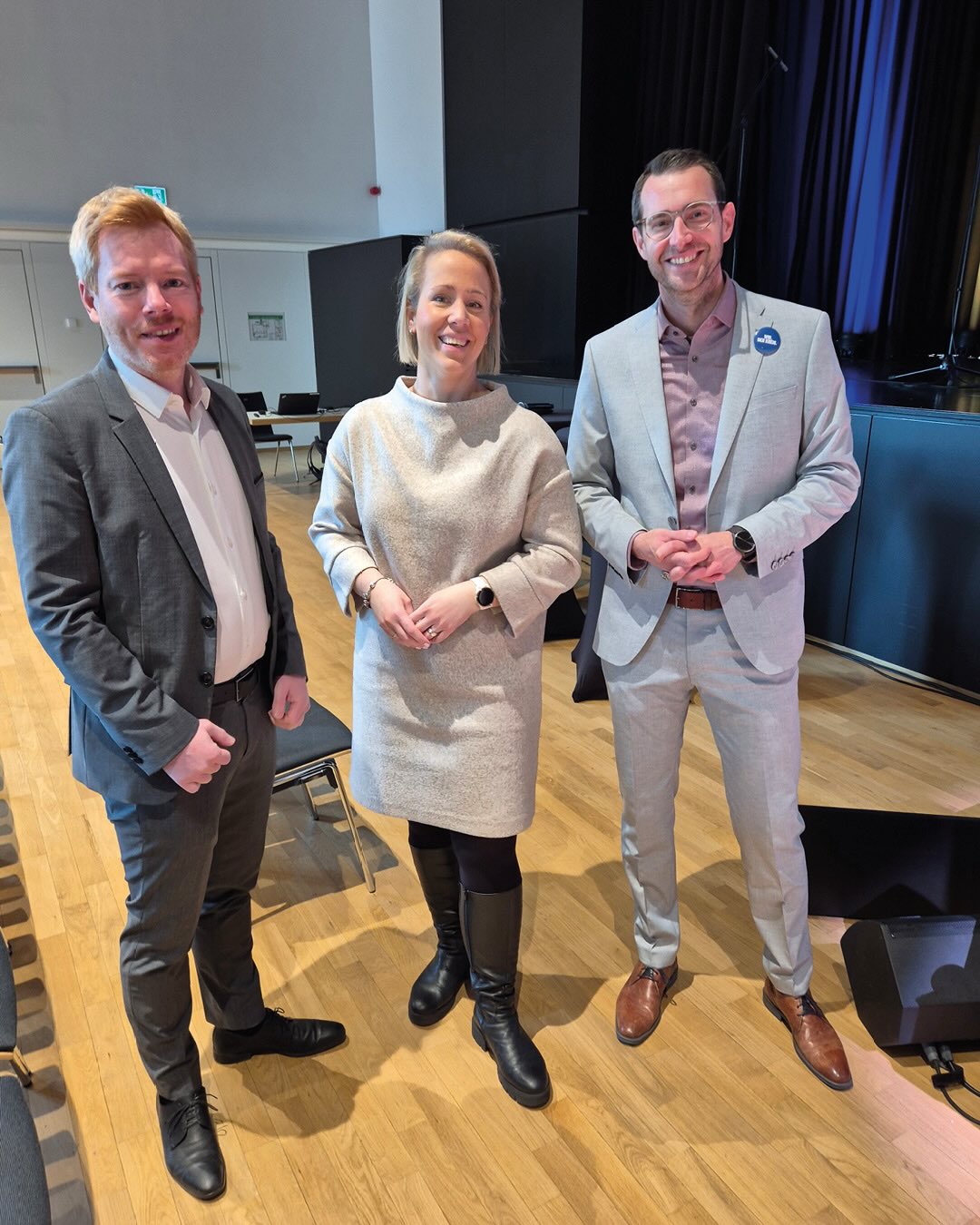 Dieser Alternativtext wurde KI-generiert: Drei Personen stehen in einem Raum mit Stühlen und einer Bühne. Alle tragen formelle Kleidung und lächeln in die Kamera.