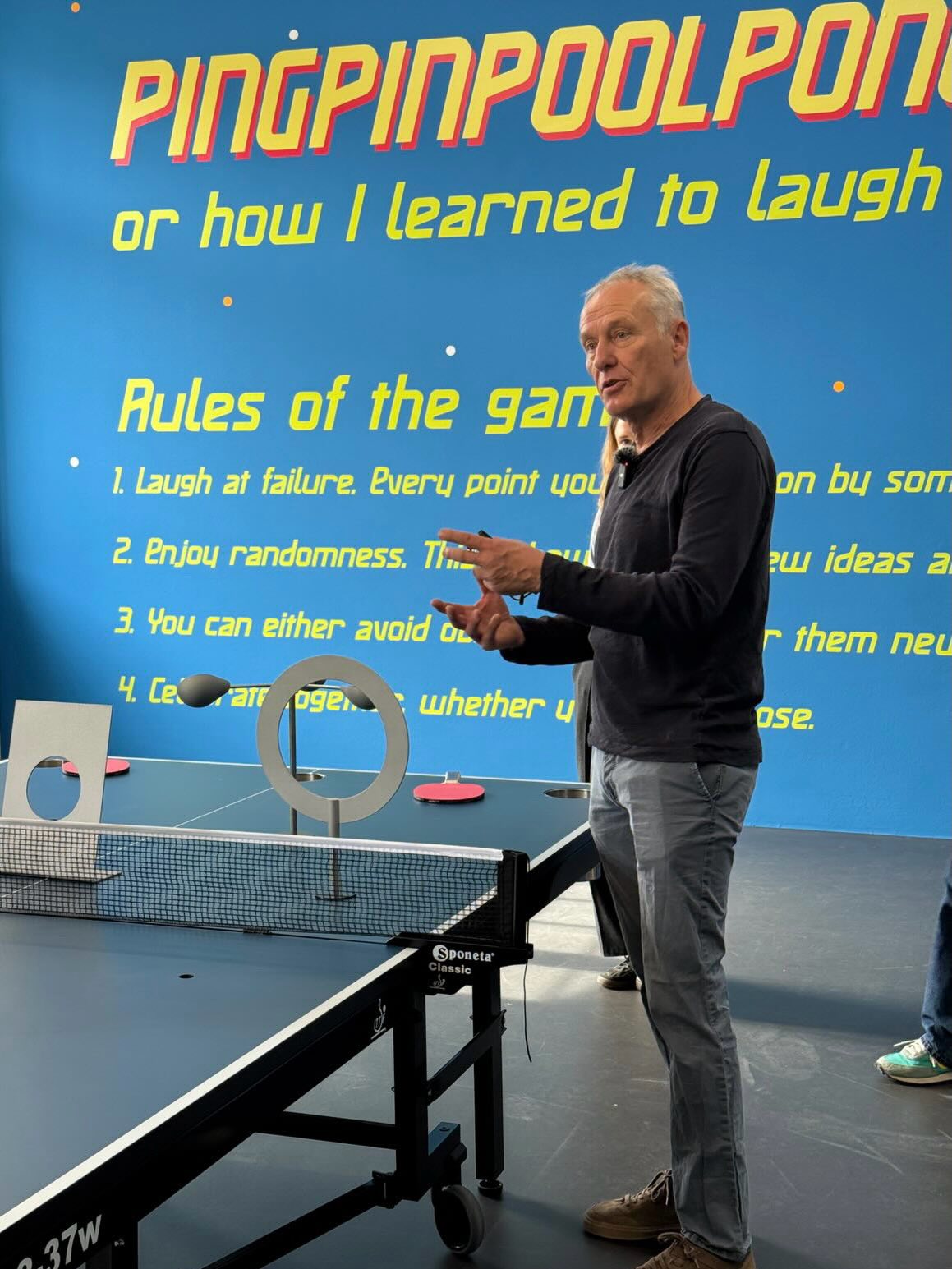 Dieser Alternativtext wurde KI-generiert: Ein Farbfoto zeigt eine Person, die auf oder neben einem Tischtennisspielgerät steht. Im Hintergrund befindet sich ein Plakat mit Text und Regeln.