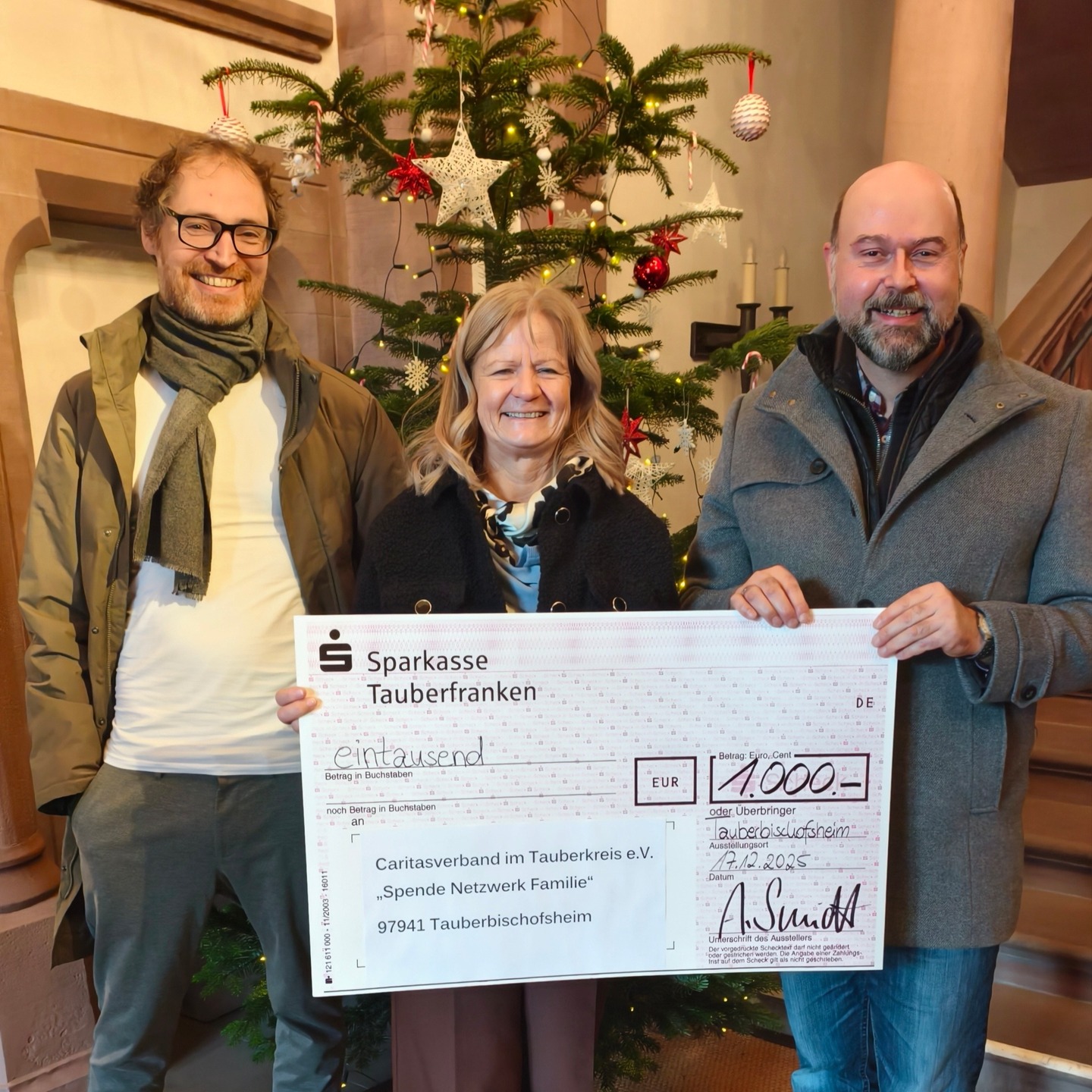 Dieser Alternativtext wurde KI-generiert: Zwei Personen halten einen Scheck über tausend Euro vor einem geschmückten Weihnachtsbaum. Im Hintergrund ist ein Spendenaufruf für ein Netzwerk in Tauberbischofsheim zu sehen.