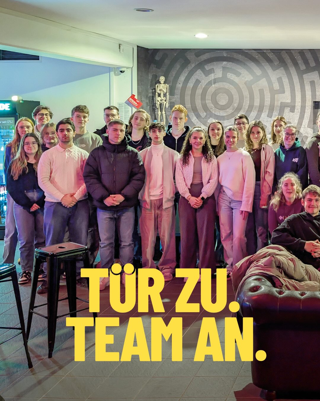 Dieser Alternativtext wurde KI-generiert: Eine Gruppe von Personen steht in einem Innenraum zusammen. Im Vordergrund befindet sich ein Banner mit Aufschrift.