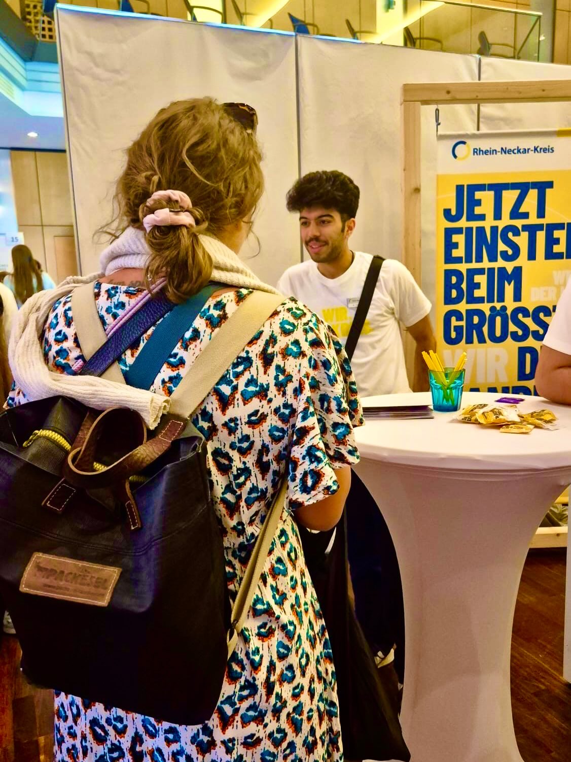 Eine Person mit einem großen schwarzen Rucksack steht mit dem Rücken zur Kamera und unterhält sich mit einer Person hinter einem weißen Hochtisch. Auf dem Tisch liegen Informationsmaterialien und ein Becher mit Stiften. Rechts im Hintergrund ist ein gelbes Banner mit dem Text „Rhein-Neckar-Kreis“ und „JETZT EINSTEIGEN BEIM GRÖSSTEN“ zu sehen. (KI-Alternativtext)