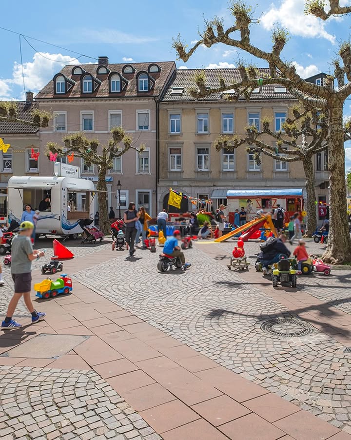 Auf einem sonnigen, gepflasterten Platz fahren Kinder auf Spielzeugautos und Roller, während Erwachsene sie beaufsichtigen. Im Hintergrund stehen mehrstöckige Gebäude, Bäume mit bunten Dekorationen sowie ein weißer und ein blauer Food-Truck. (KI-Alternativtext)