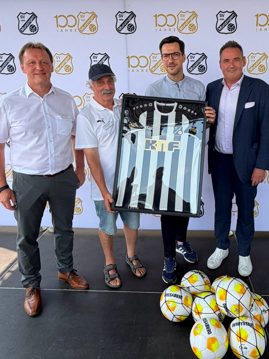 Vier Personen stehen vor einem Hintergrundbanner mit dem Logo "SV Munzingen" und der Aufschrift "100 Jahre". Zwei Personen in der Mitte halten gemeinsam ein gerahmtes, schwarz-weiß gestreiftes Fußballtrikot mit der Aufschrift "KIF". Auf dem Boden im Vordergrund liegen mehrere weiße Fußball. (KI-Alternativtext)