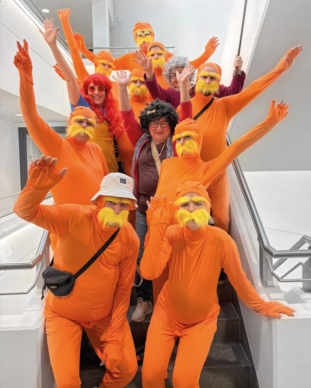 Dieser Alternativtext wurde KI-generiert: Eine Gruppe von Personen in orangefarbenen Ganzkörperanzügen und Accessoires posiert auf einer Treppe. Einige tragen Bärte und Brillen.