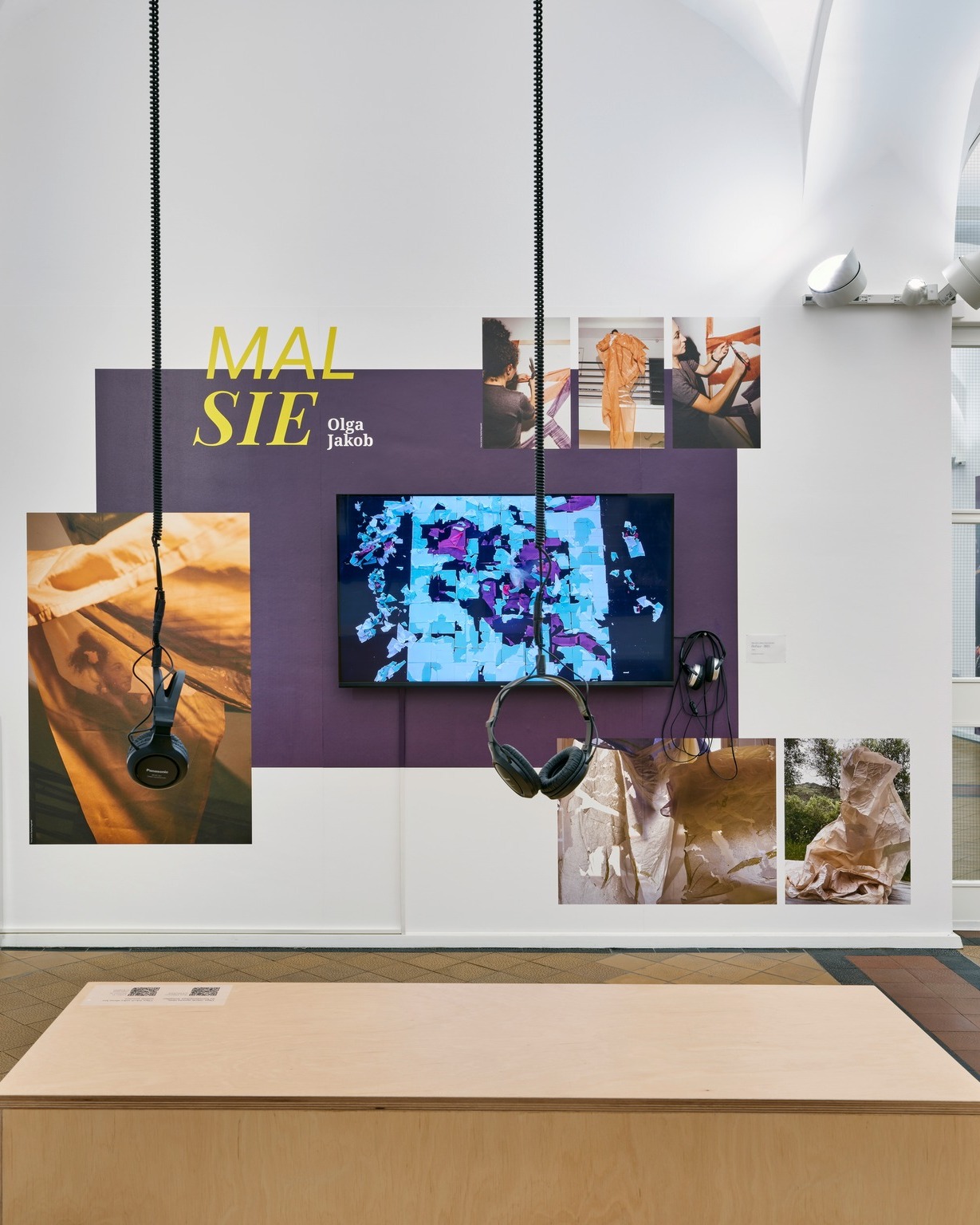 Dieser Alternativtext wurde KI-generiert: Eine Innenansicht einer Kunstausstellung mit mehreren Bildern an einer Wand und Kopfhörern davor. Das Bild ist farbig und zeigt verschiedene künstlerische Arbeiten.