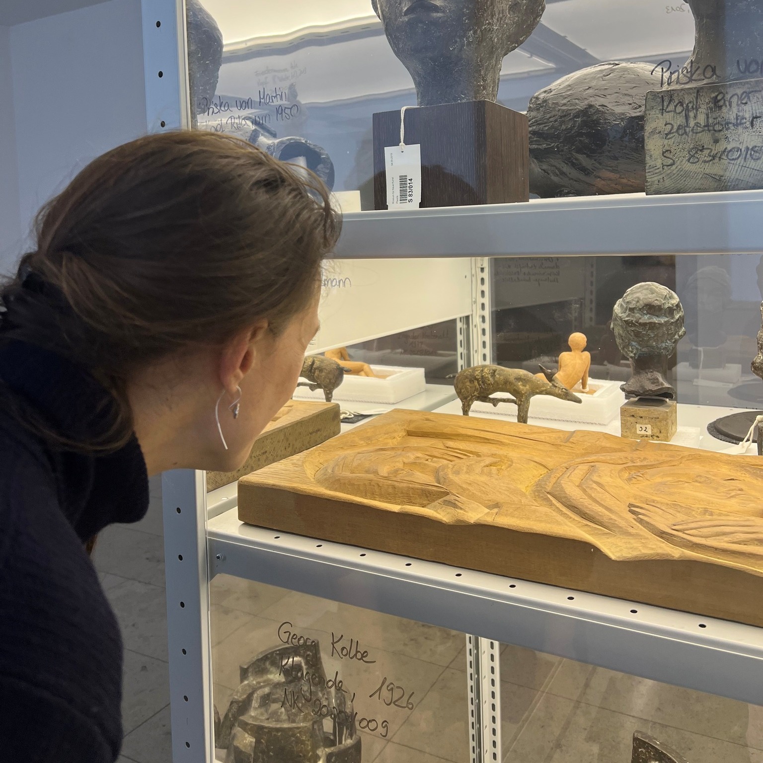 Dieser Alternativtext wurde KI-generiert: Eine Person betrachtet Ausstellungsstücke in einer Vitrine. Zu sehen sind verschiedene Skulpturen und Objekte auf mehreren Ebenen.