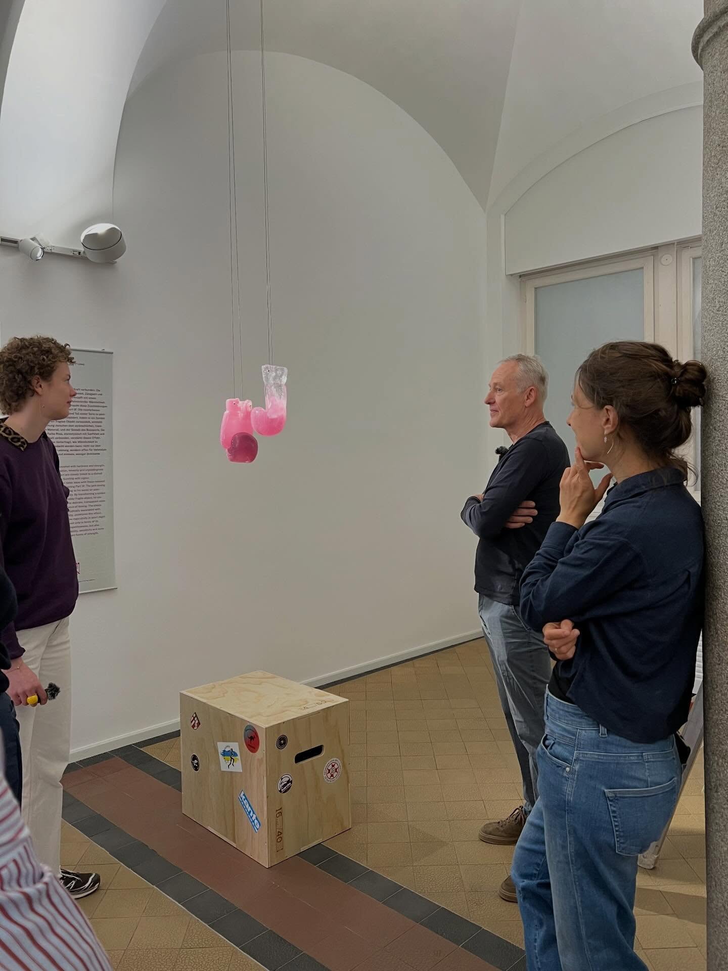 Dieser Alternativtext wurde KI-generiert: Eine Gruppe von Personen steht in einem Innenraum vor einer weißen Wand und betrachtet eine Installation, die von der Decke hängt. Im Vordergrund steht eine Holzkonstruktion mit Aufklebern auf einem Karton.