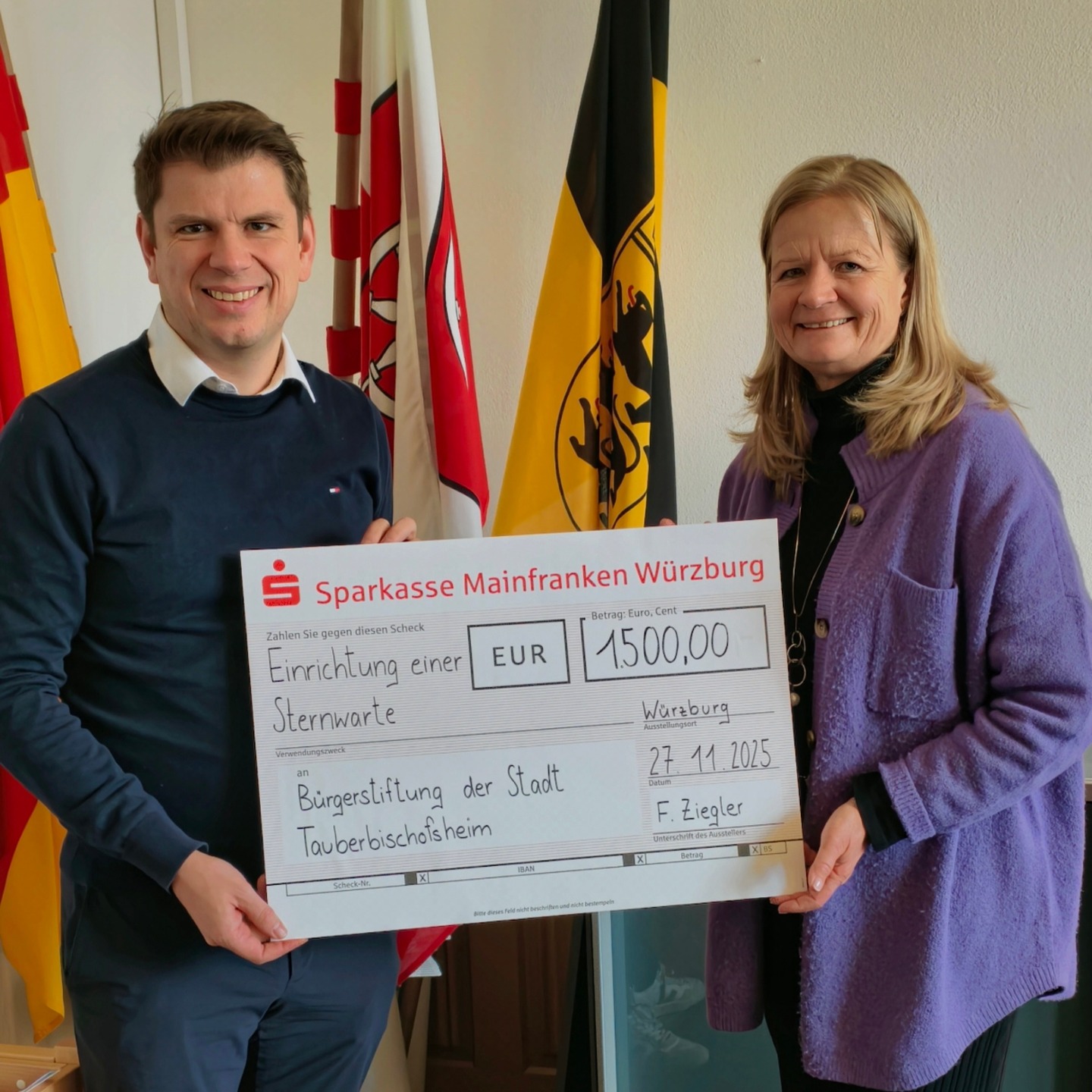 Dieser Alternativtext wurde KI-generiert: Zwei Personen posieren mit einem großen Scheck. Der Scheck ist von einer Sparkasse und wird an eine Bürgerstiftung überreicht.