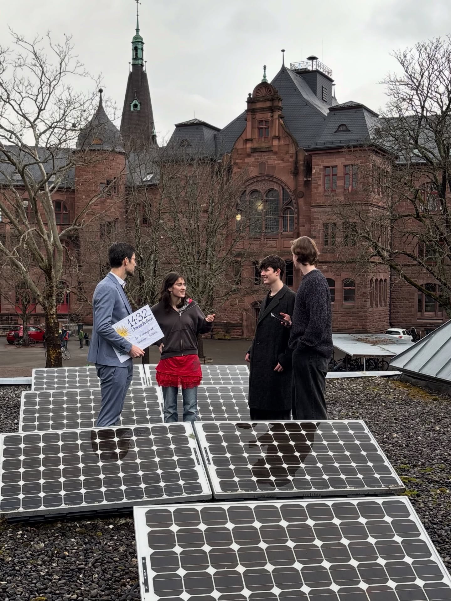 Dieser Alternativtext wurde KI-generiert: Mehrere Personen stehen vor einem historischen Gebäude mit roten Backsteinen und einer markanten Turmspitze. Im Vordergrund befinden sich Solarpanels und die Menschen halten Schilder in den Händen.