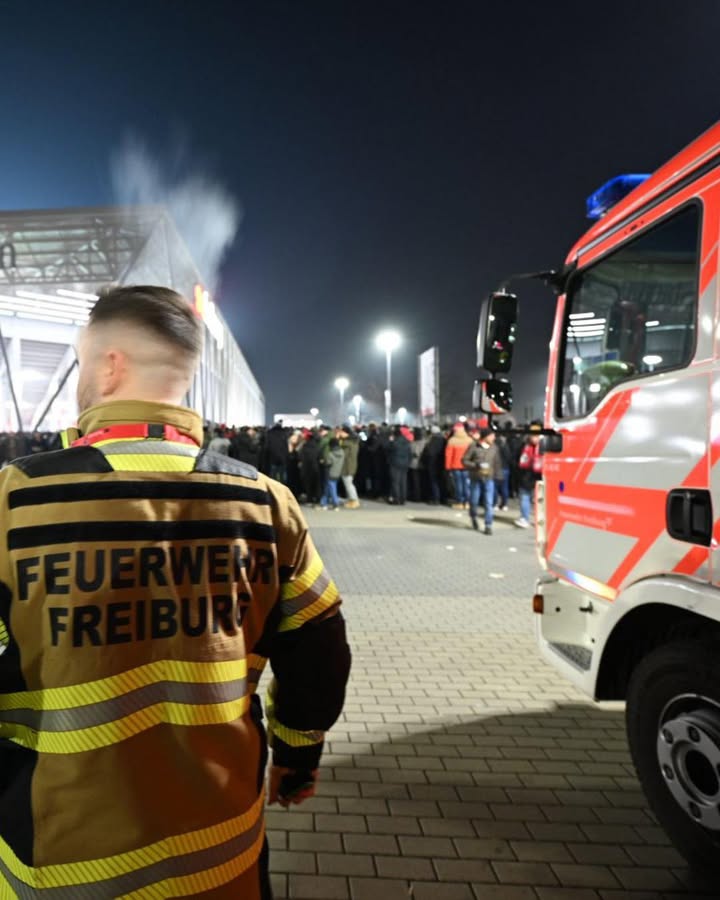 Dieser Alternativtext wurde KI-generiert: Eine Person in einer reflektierenden Jacke steht mit dem Rücken zum Betrachter vor einem roten Einsatzfahrzeug. Im Hintergrund sind Menschen und ein Gebäude zu erkennen.