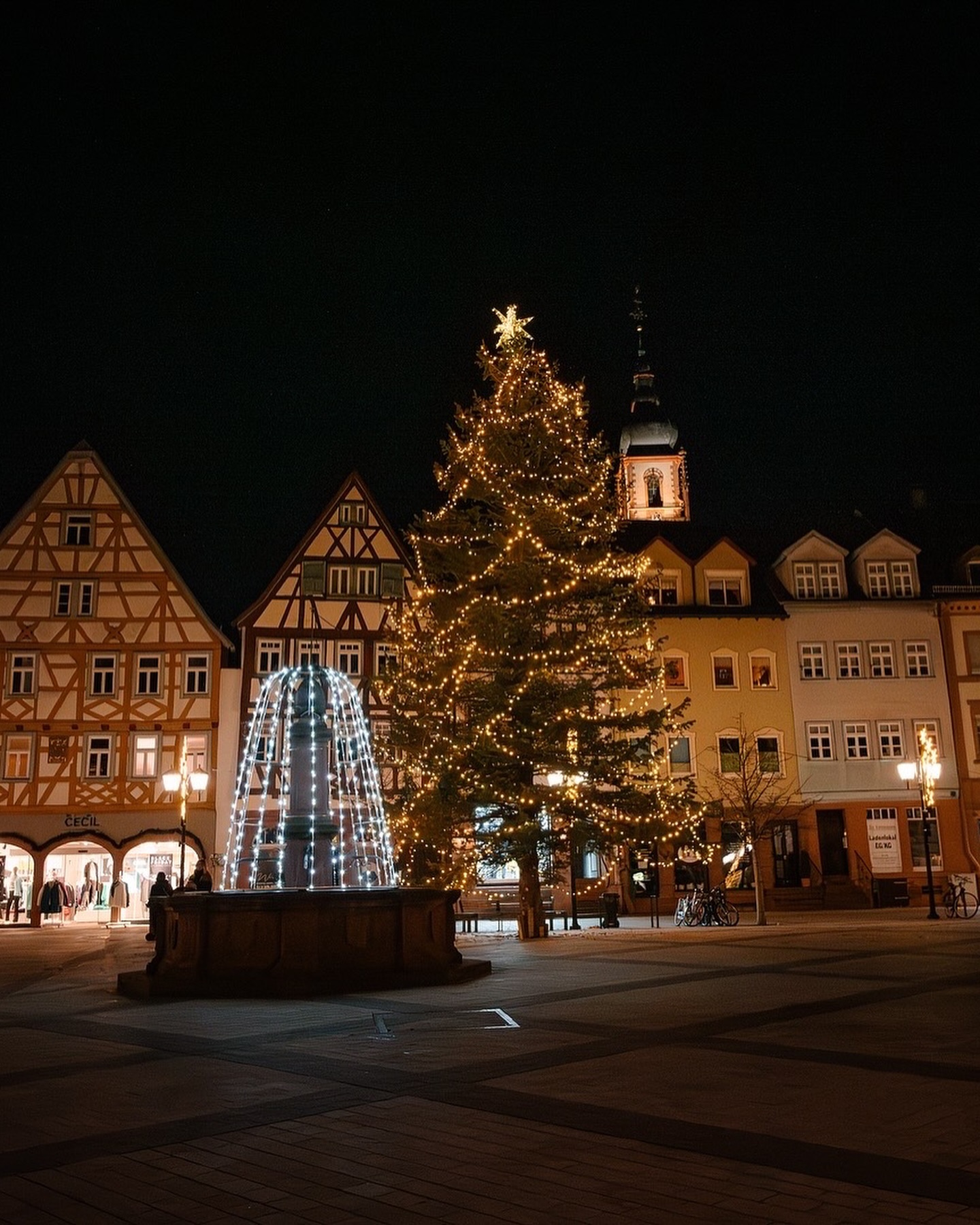 Ein beleuchteter Weihnachtsbaum steht auf einem Platz zwischen Fachwerkhäusern. Eine Brunnenanlage ist ebenfalls weihnachtlich geschmückt.