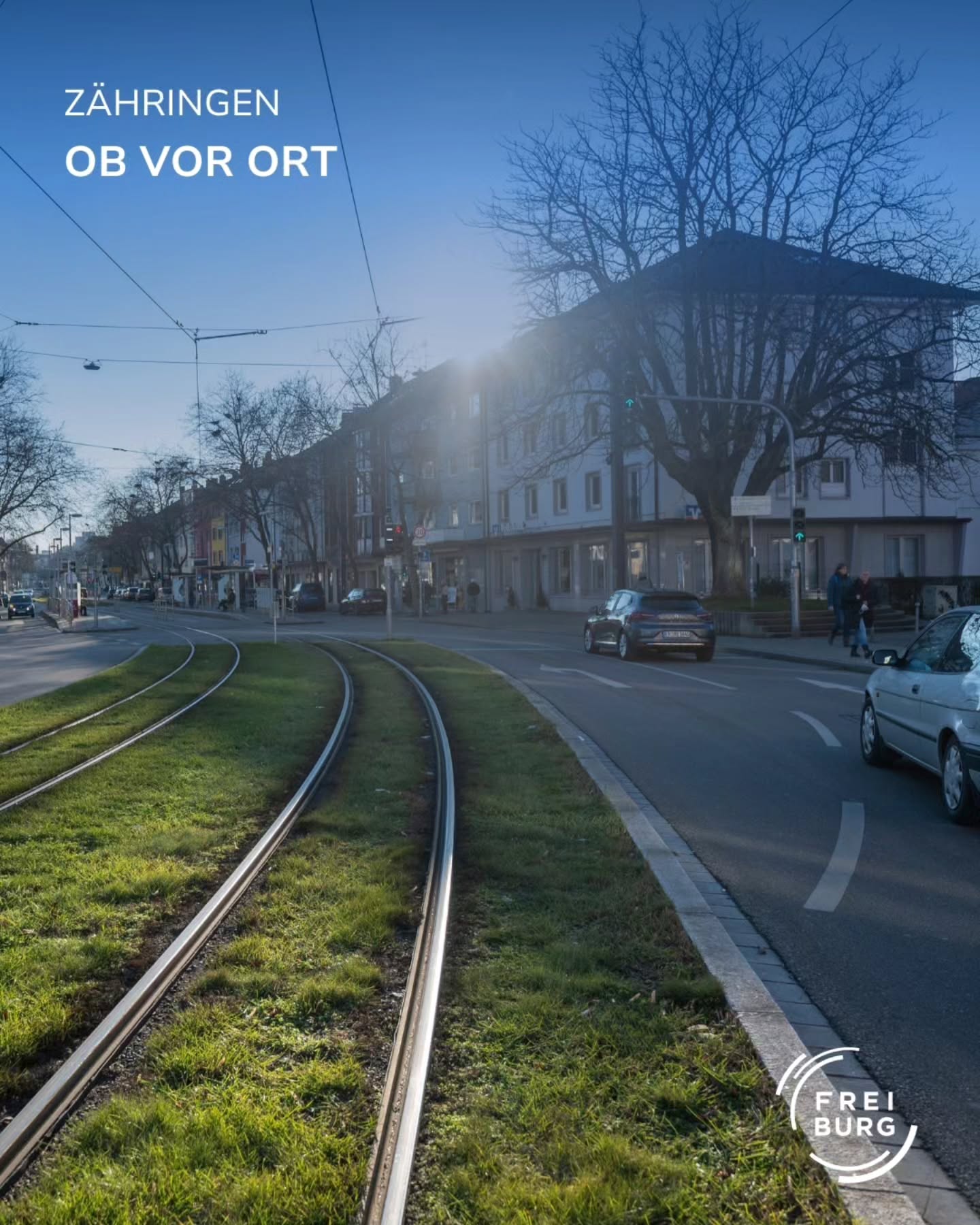 Dieser Alternativtext wurde KI-generiert: Eine Straßenansicht mit Straßenbahnschienen im Vordergrund und Gebäuden sowie Bäumen im Hintergrund. Das Bild ist farbig und zeigt eine städtische Szene.