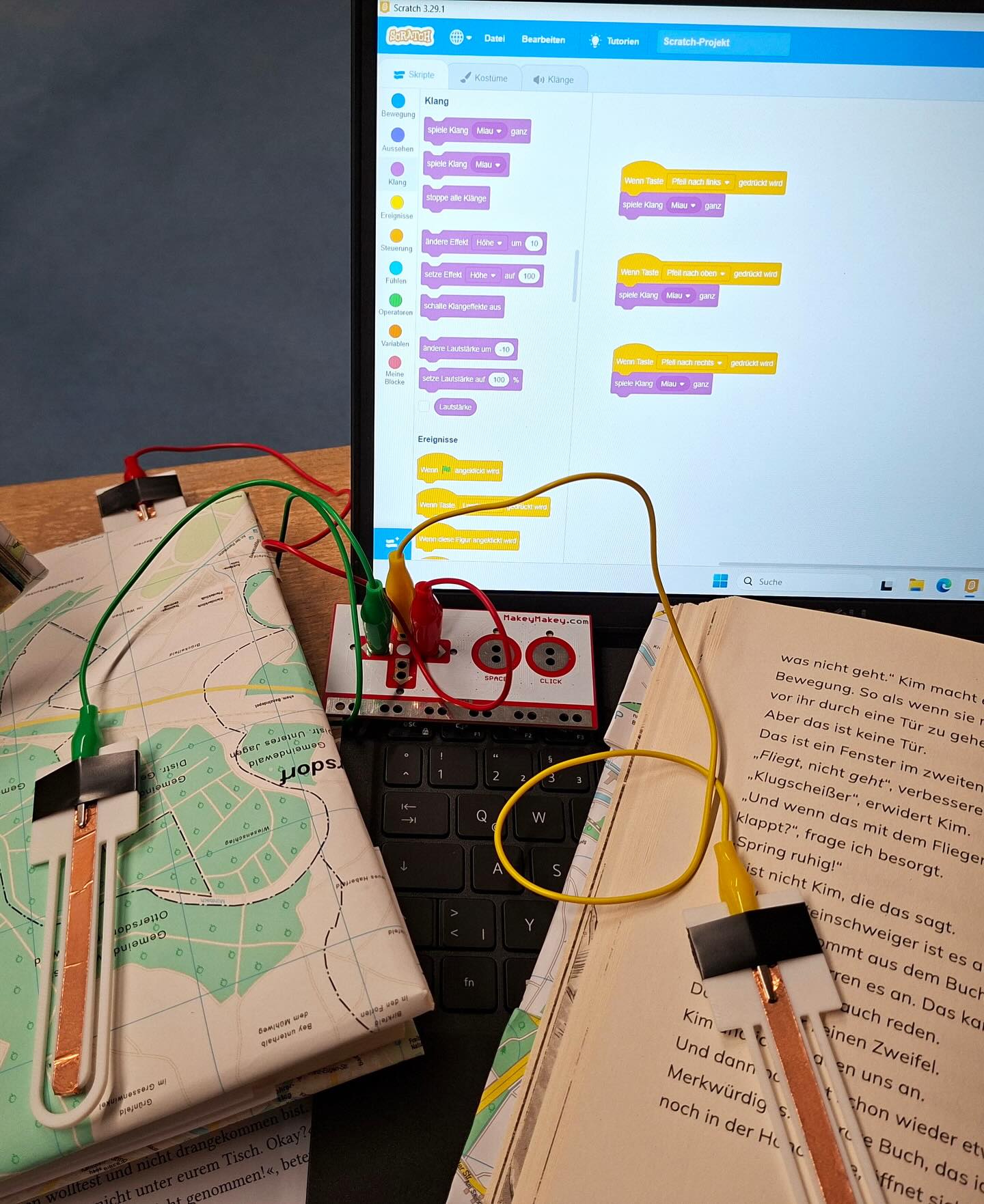 Dieser Alternativtext wurde KI-generiert: Ein farbiges Foto zeigt einen Laptop, darauf ein geöffnetes Computerprogramm mit vielen grafischen Elementen. Neben dem Laptop liegen ein Buch, eine Brille und weitere Gegenstände auf einer Oberfläche.