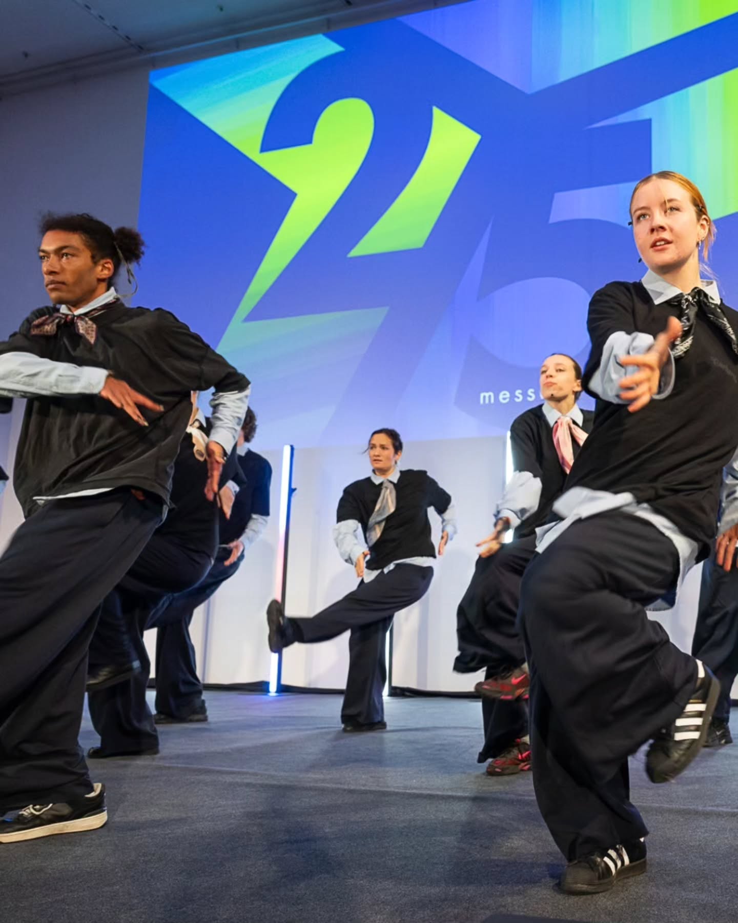 Dieser Alternativtext wurde KI-generiert: Mehrere Personen in formeller Kleidung mit lockeren Elementen führen eine synchronisierte Tanzperformance auf einer Bühne auf. Im Hintergrund ist ein großes, farbiges Logo und ein Schriftzug zu sehen.