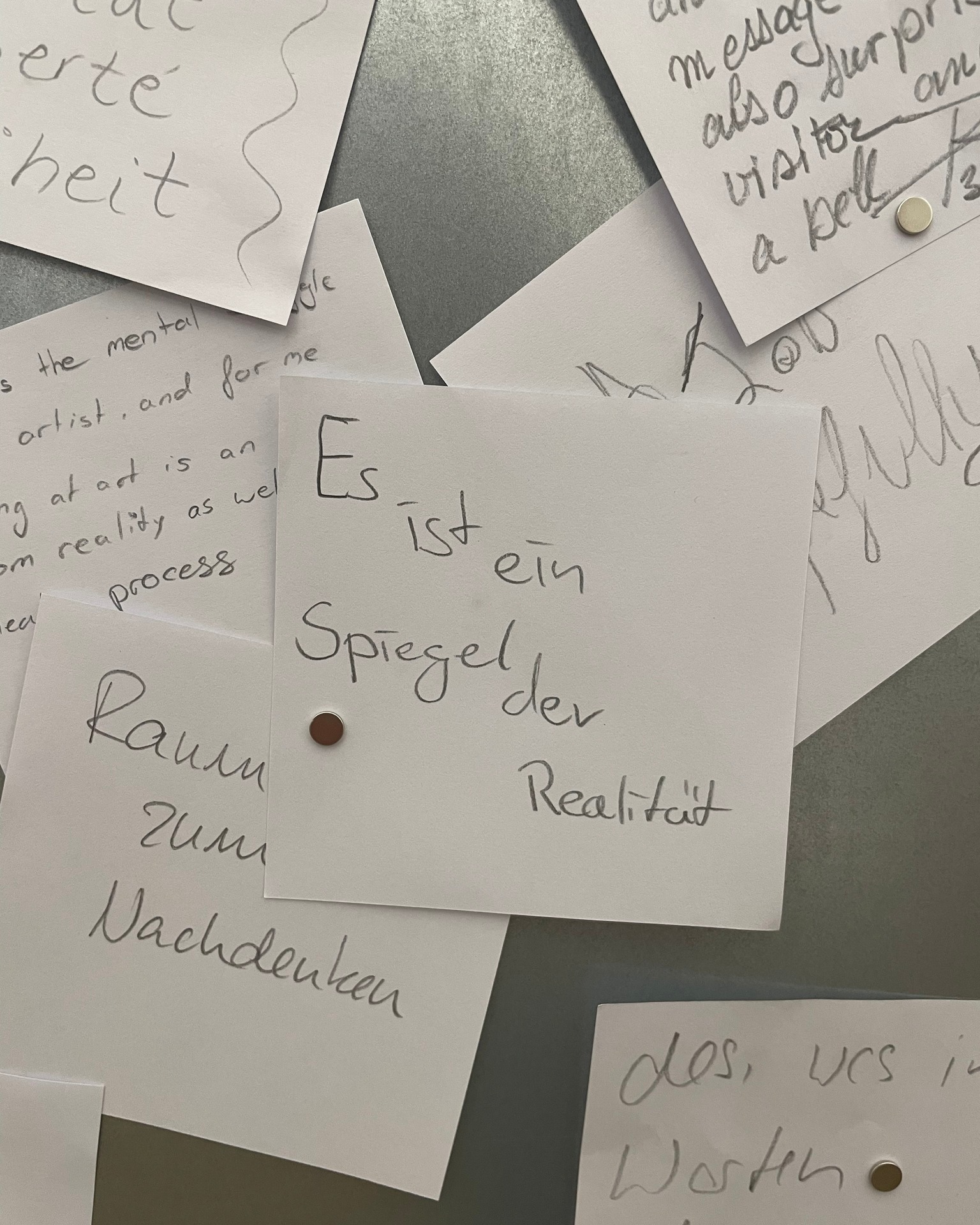 Dieser Alternativtext wurde KI-generiert: Mehrere beschriftete Zettel liegen lose auf einer grauen Oberfläche. Die Handschrift und die Texte scheinen philosophischer Natur zu sein.