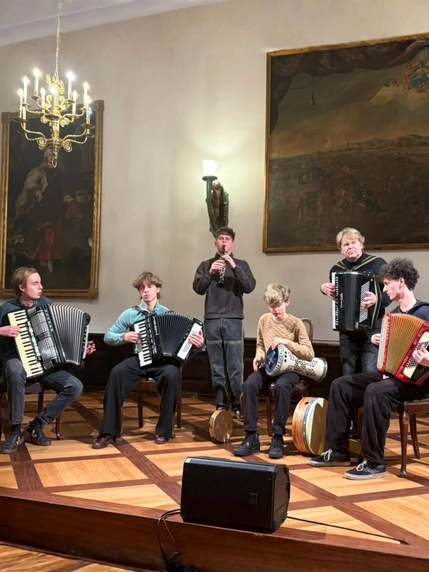 Dieser Alternativtext wurde KI-generiert: Eine Gruppe von Personen spielt verschiedene Instrumente, darunter Zieharmonikas und eine Trommel, in einem Raum mit kunstvollen Gemälden und einem Kronleuchter. Der Boden ist parkettartig verlegt.