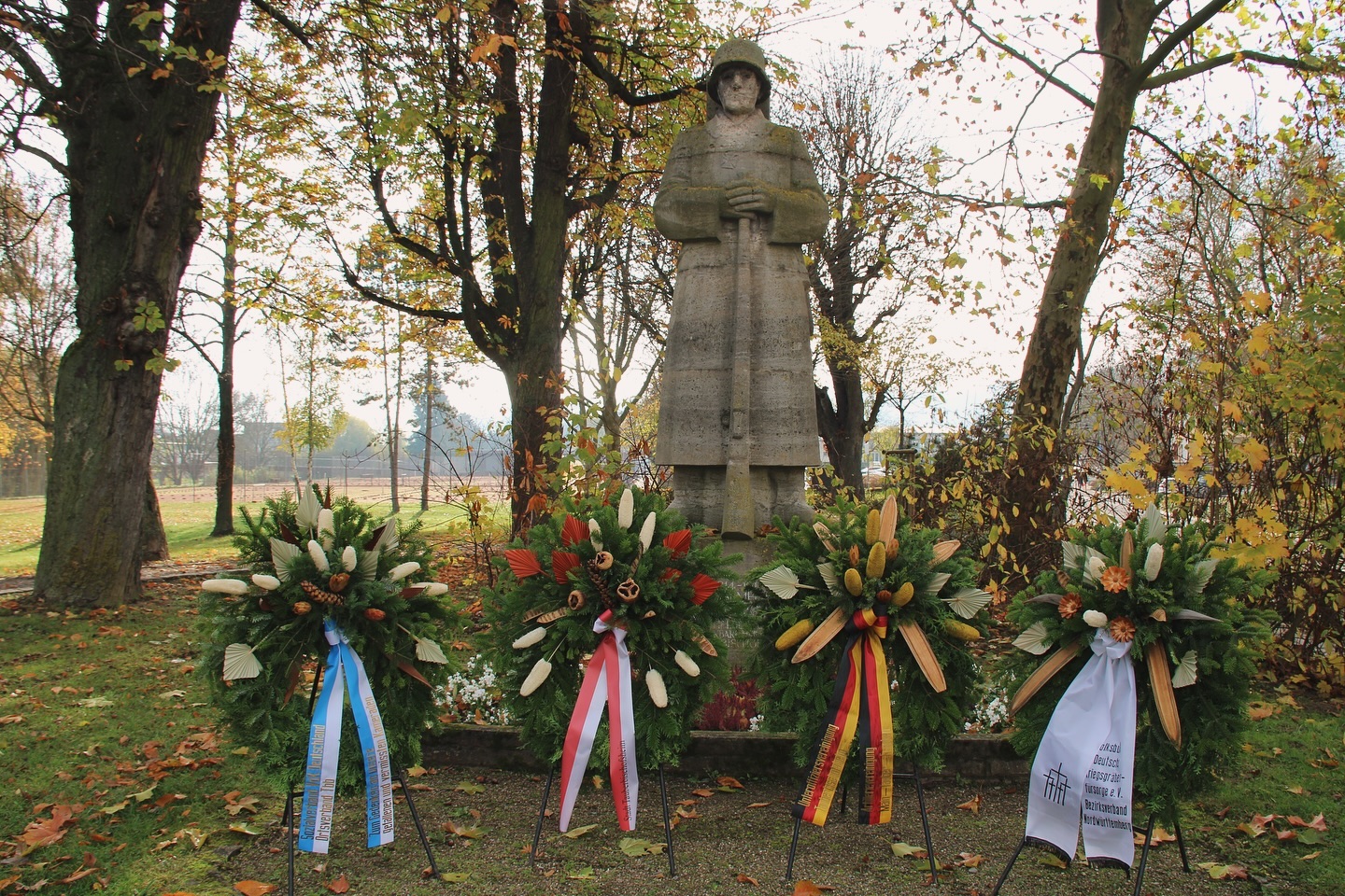 Ein Soldatenstandbild steht inmitten von drei Kränzen in einem herbstlichen Park. Die Kränze sind mit Schleifen versehen.
