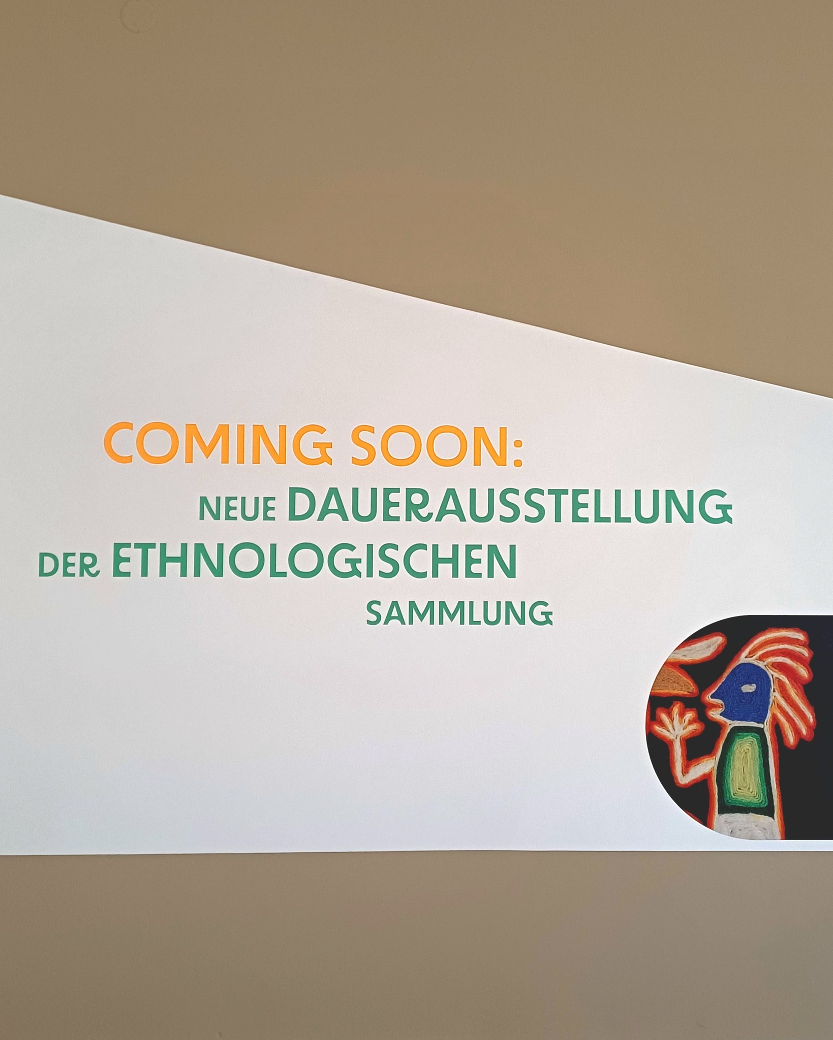 Ein weißes Schild an einer Wand trägt die Aufschrift "COMING SOON: NEUE DAUERAUSSTELLUNG DER ETHNOLOGISCHEN SAMMLUNG". Unten rechts ist ein rundes Motiv mit einer stilisierten Figur in Blau und Rot zu sehen. (KI-Alternativtext)