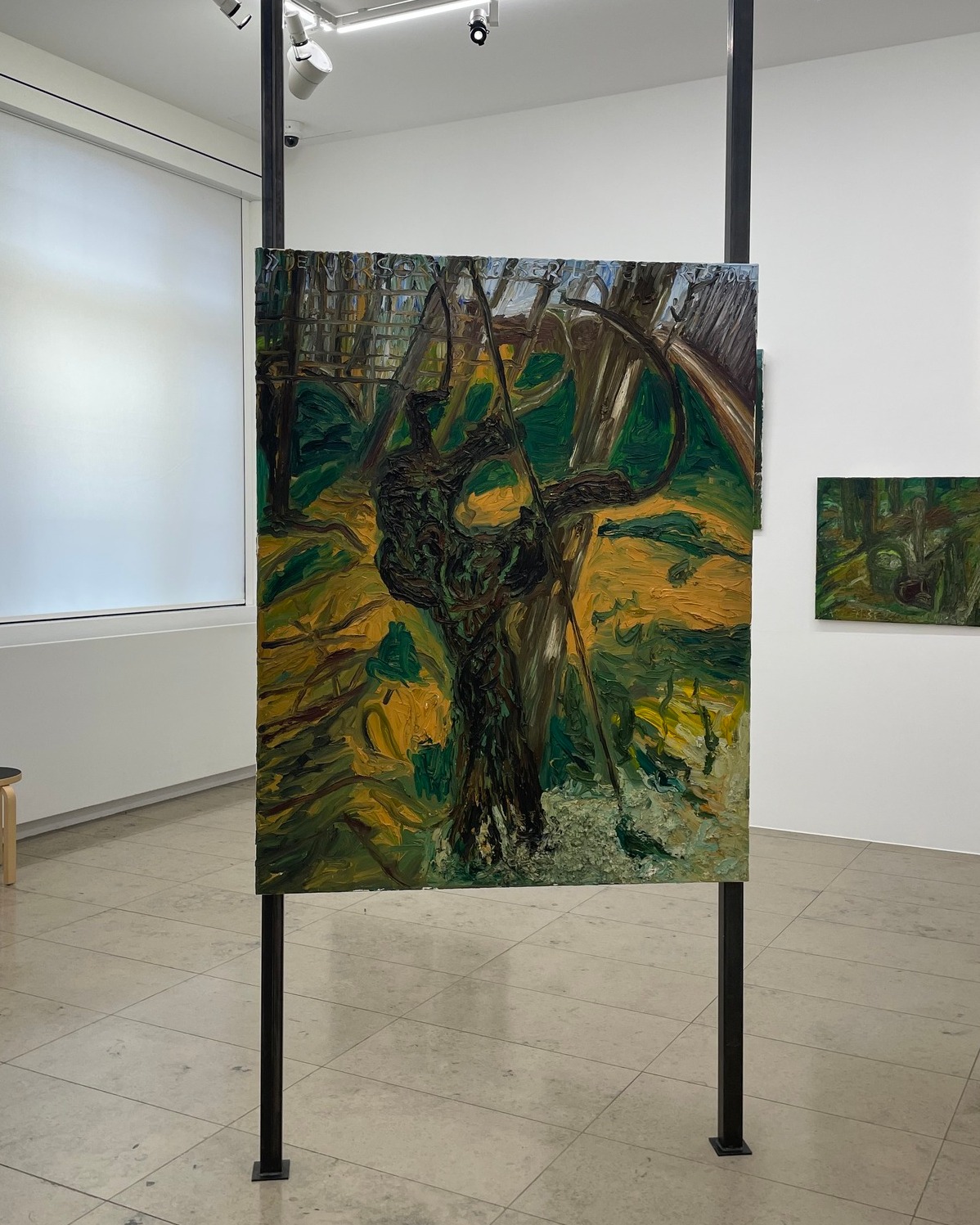 Dieser Alternativtext wurde KI-generiert: Ein Raum mit Kunstwerken an den Wänden. Im Fokus steht ein großes, farbenfrohes Gemälde mit Baumdarstellungen, das auf einem schwarzen Stahlgestell steht.