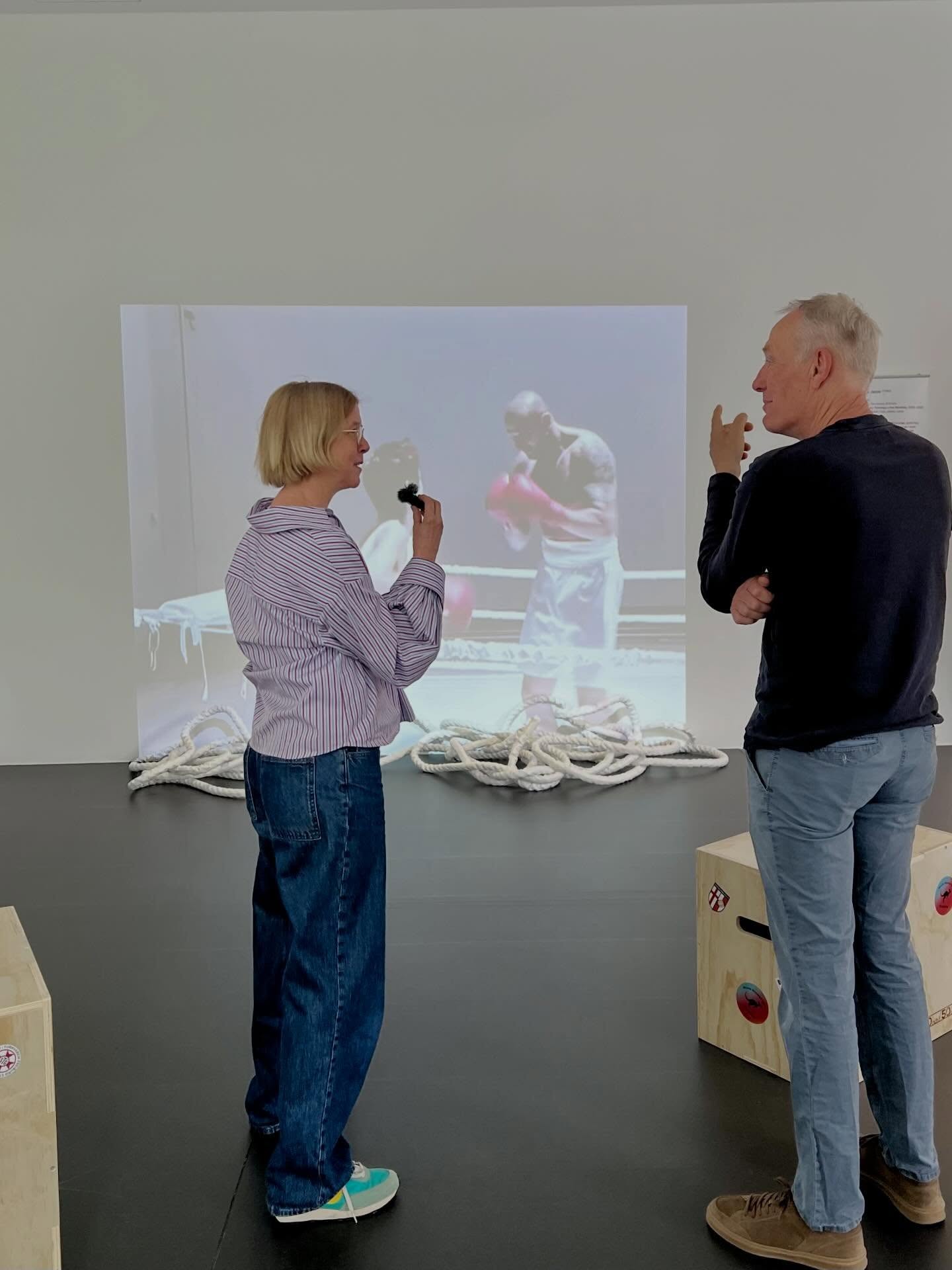 Dieser Alternativtext wurde KI-generiert: Zwei Personen betrachten ein Videoprojektion in einem hellen Raum. Im Vordergrund befindet sich ein Seil vor einem schwarzen Hintergrund.