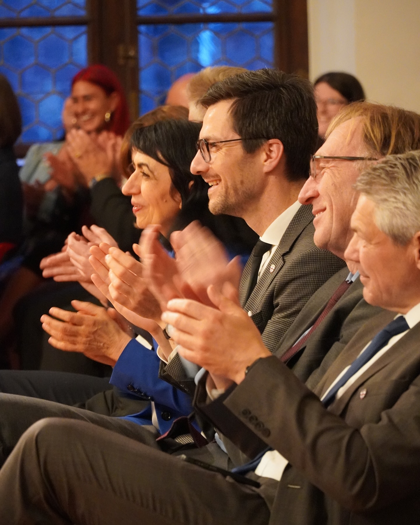 Eine Gruppe von Menschen applaudiert. Sie sitzen in einem Raum und tragen formelle Kleidung.