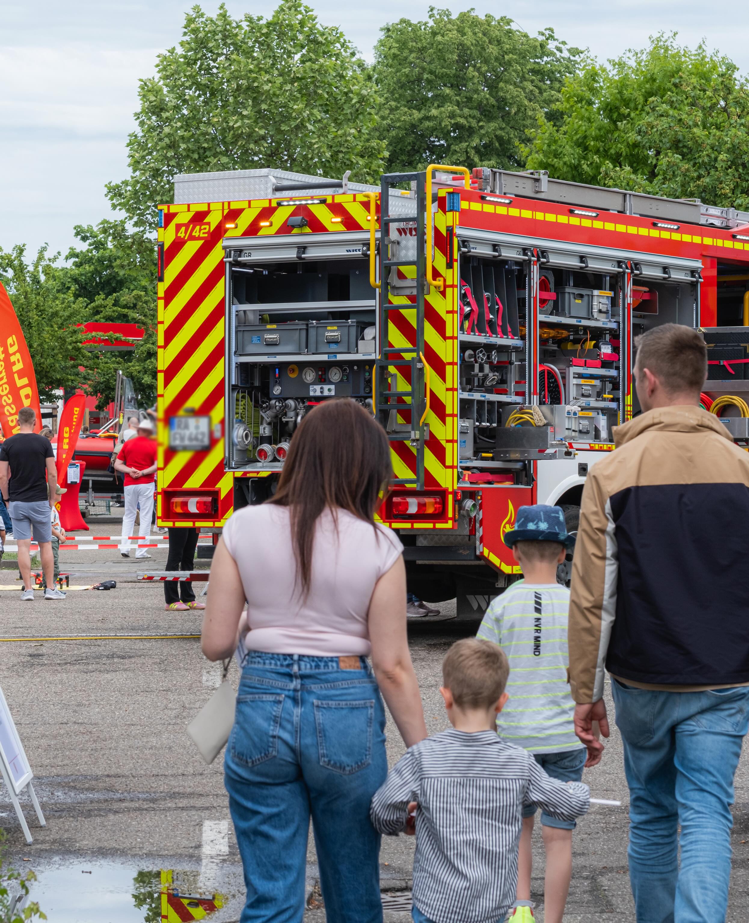 Dieser Alternativtext wurde KI-generiert: Ein rotes Feuerwehrauto mit gelben Markierungen steht auf einer Straße. Mehrere Personen betrachten das Fahrzeug von hinten, während sie auf dem Bürgersteig stehen.