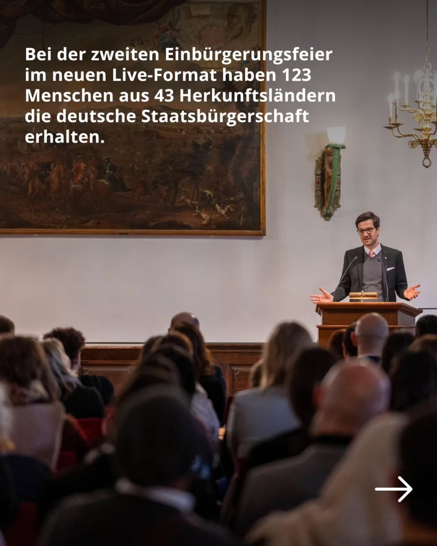 Dieser Alternativtext wurde KI-generiert: Ein Innenraum mit einer Person, die vor einer Gruppe von Menschen an einem Rednerpult spricht. Im Hintergrund befindet sich ein großes Gemälde und eine dekorative Wandgestaltung.