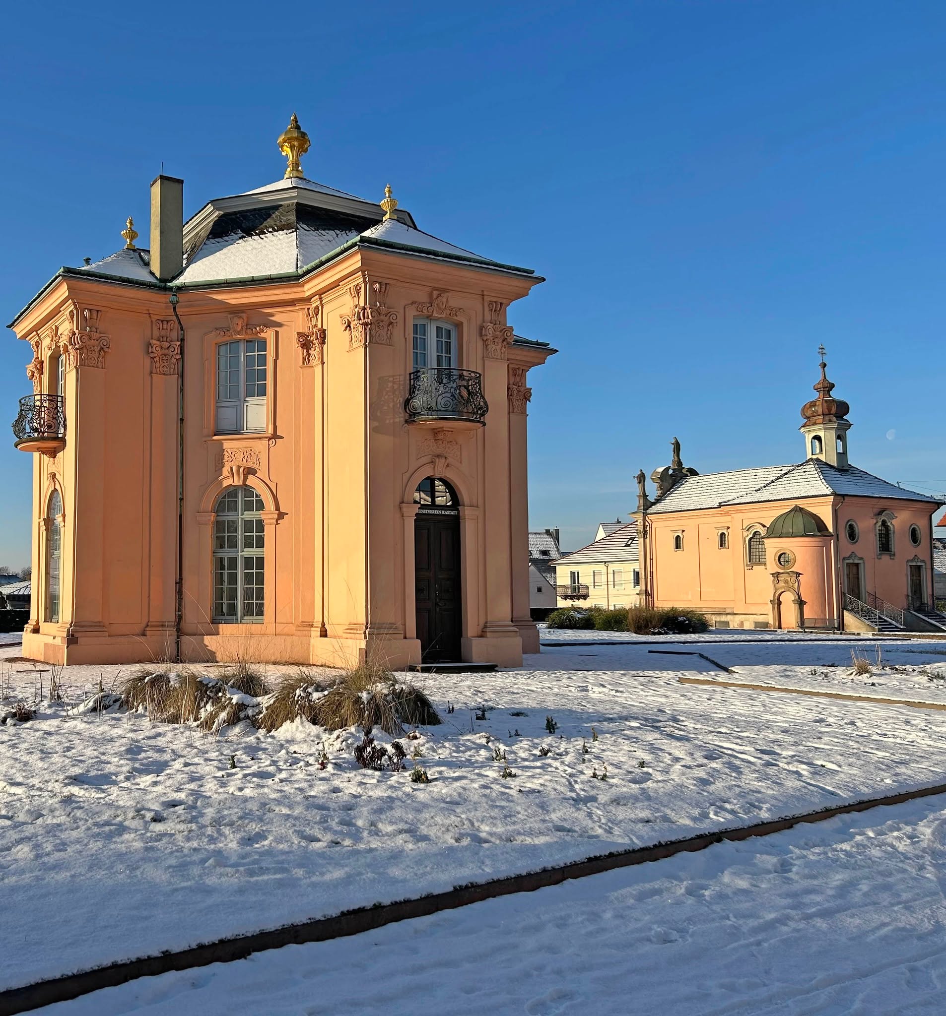 Dieser Alternativtext wurde KI-generiert: Ein helles, pastellfarbenes Gebäude mit Kuppel steht auf einer schneebedeckten Fläche. Im Hintergrund ist ein weiteres, kleineres Gebäude zu sehen, vor einem klaren, blauen Himmel.