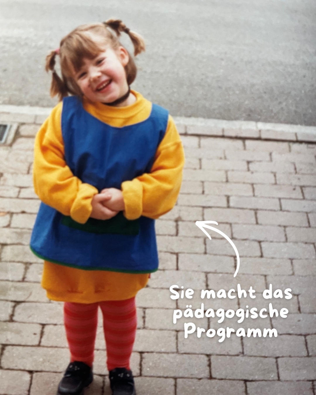 Dieser Alternativtext wurde KI-generiert: Ein Schwarzweißfoto zeigt eine Person, die auf einem gepflasterten Weg steht und lächelt. Die Person trägt einen Overall und rote Strumpfhosen.