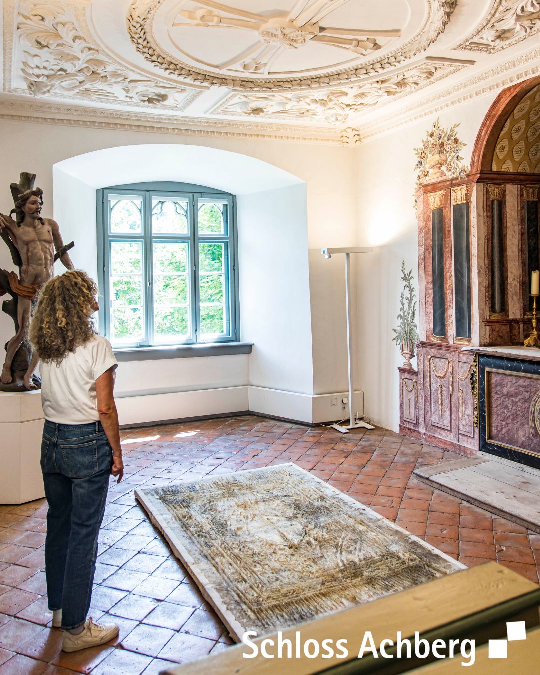 Ein Innenraum mit einem Fenster und detailreichen Verzierungen an Wänden und Decke. Eine Person steht mit dem Rücken zum Betrachter vor einem Teppich und betrachtet die Umgebung.