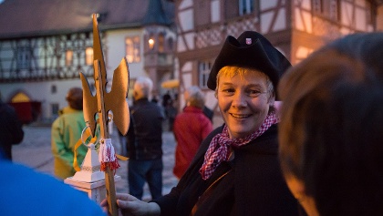 Person im historischen Kostüm hält eine Stange mit goldenem Aufsatz. Im Hintergrund ist ein Fachwerkhaus zu sehen.