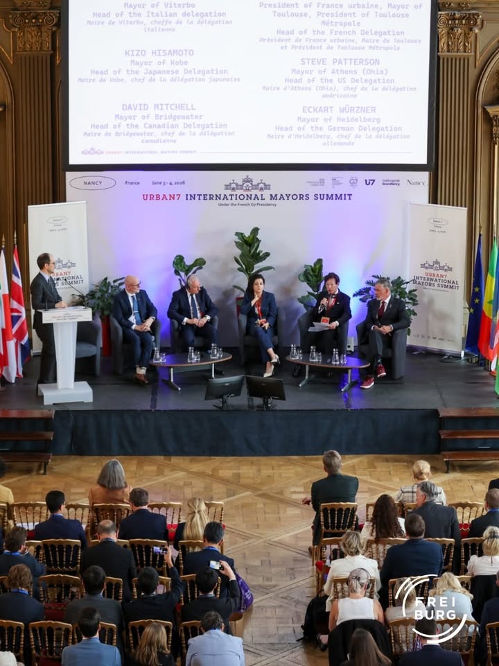 Auf einer Bühne sitzen sechs Personen in Anzügen, eine steht am Rednerpult. Hinter ihnen ein Bildschirm mit Namen und Funktionen der Delegationen sowie der Aufschrift „URBAN7 INTERNATIONAL MAYORS SUMMIT“. Im Vordergrund sitzt ein Publikum vor Holzstühlen, rechts und links stehen Flaggen und Werbebanner. (KI-Alternativtext)