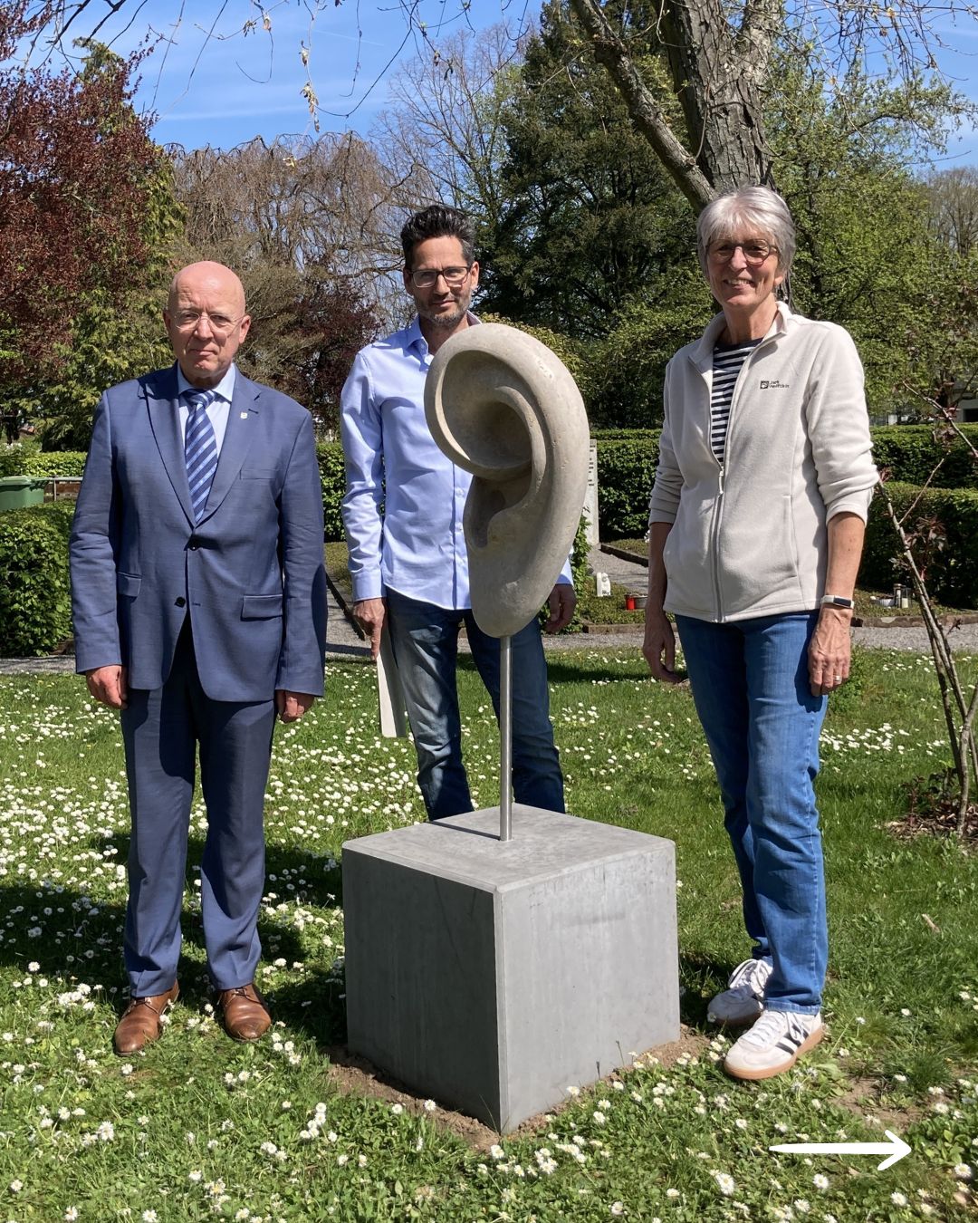 Dieser Alternativtext wurde KI-generiert: Drei Personen stehen neben einer modernen Skulptur in einem Garten. Im Hintergrund befindet sich grünes Laub und eine Wiese voller kleiner, weißer Blumen.