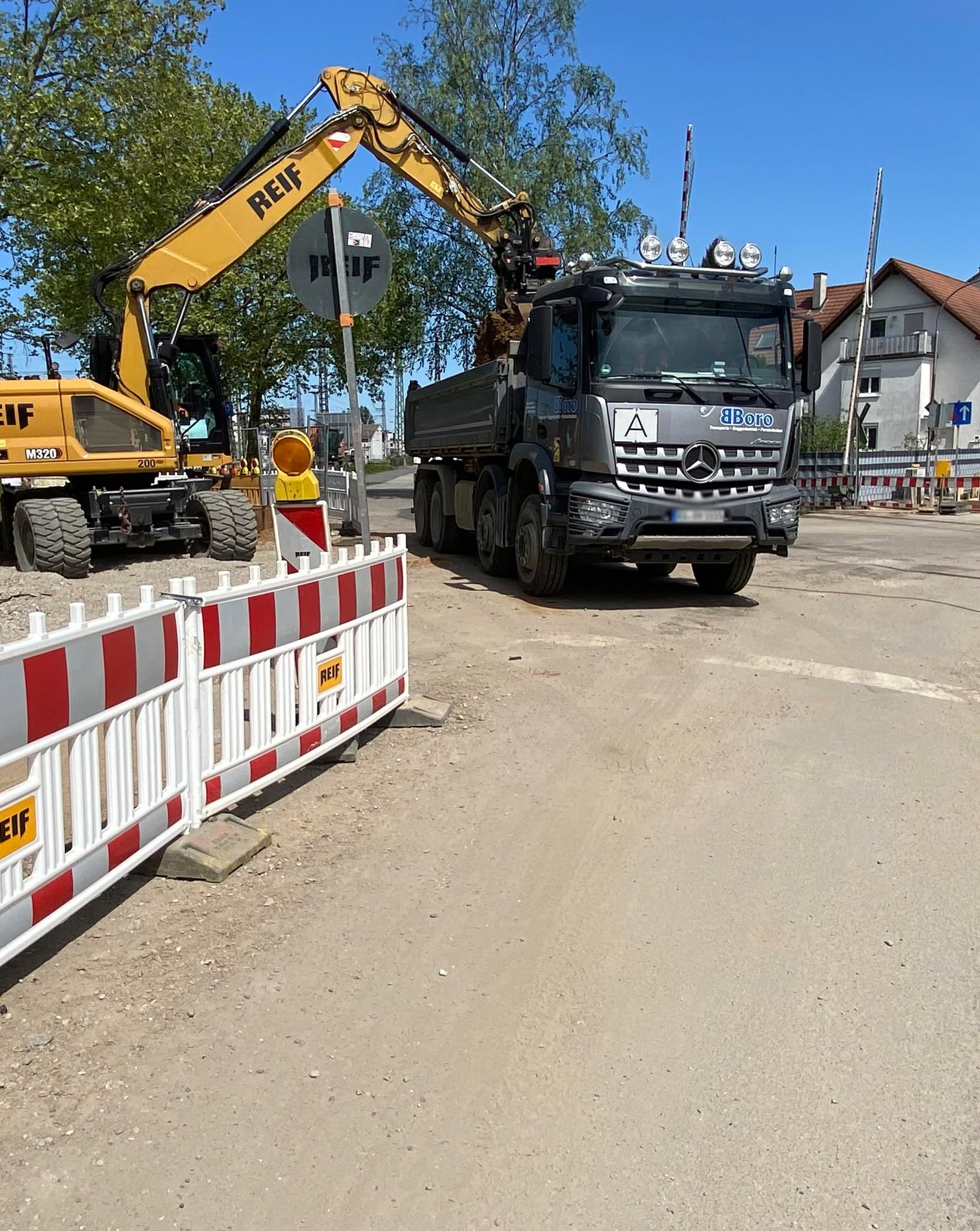 Dieser Alternativtext wurde KI-generiert: Ein Bagger und ein Lastwagen stehen auf einer Baustelle neben einer orangefarbenen Absperrung. Im Hintergrund sind Gebäude und weitere Baustellenutensilien zu sehen.