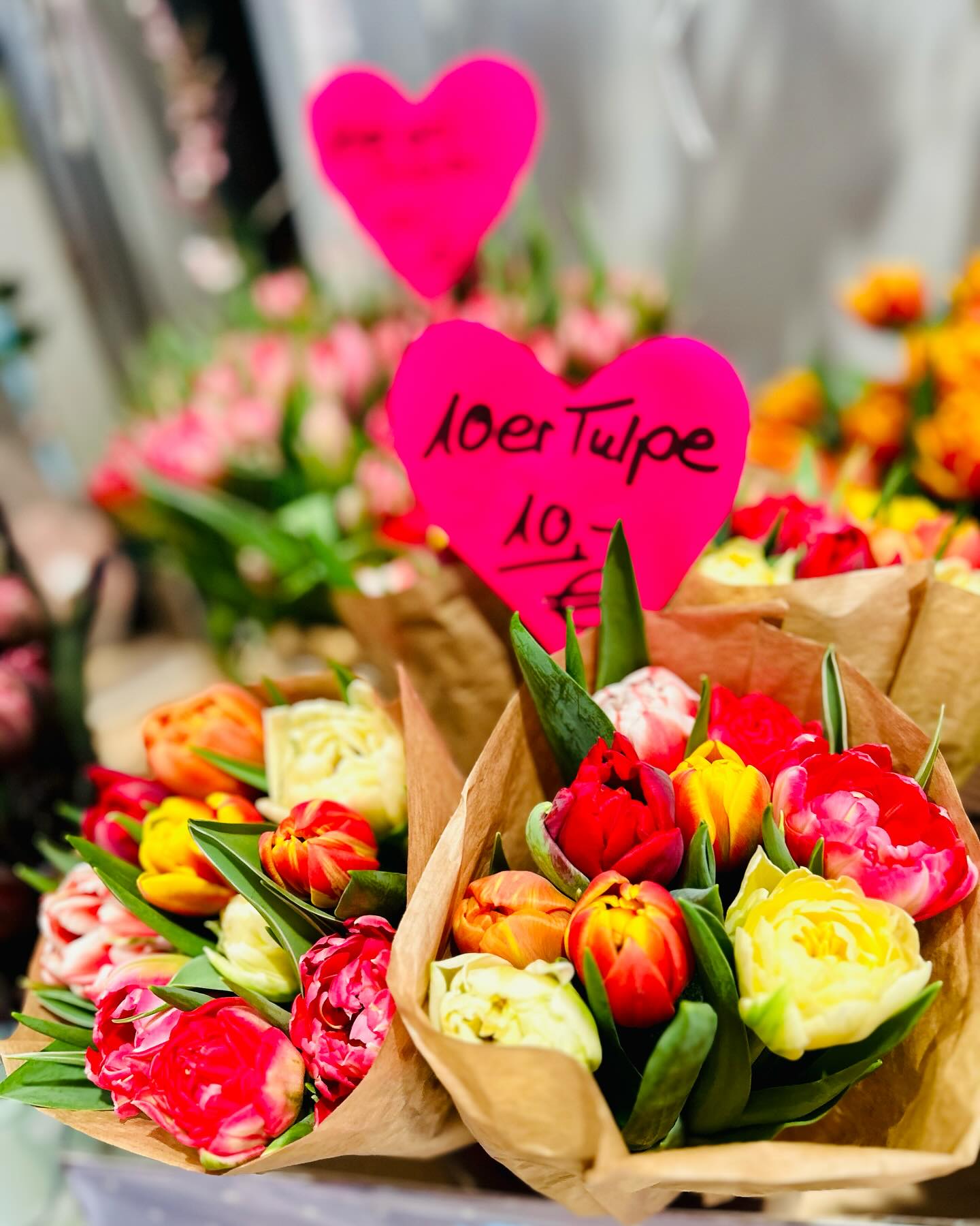 Mehrere Blumensträuße mit Tulpen in verschiedenen Farben sind in Papier verpackt. Herzen mit Preisen sind an den Sträußen befestigt.