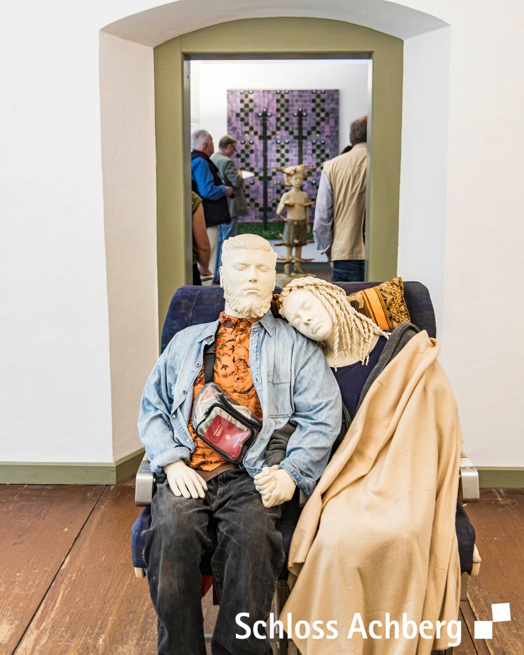 Ein Raum mit einer Installation, die menschliche Formen auf einem Sitz zeigt. Im Hintergrund sind weitere Kunstwerke und Personen zu sehen.