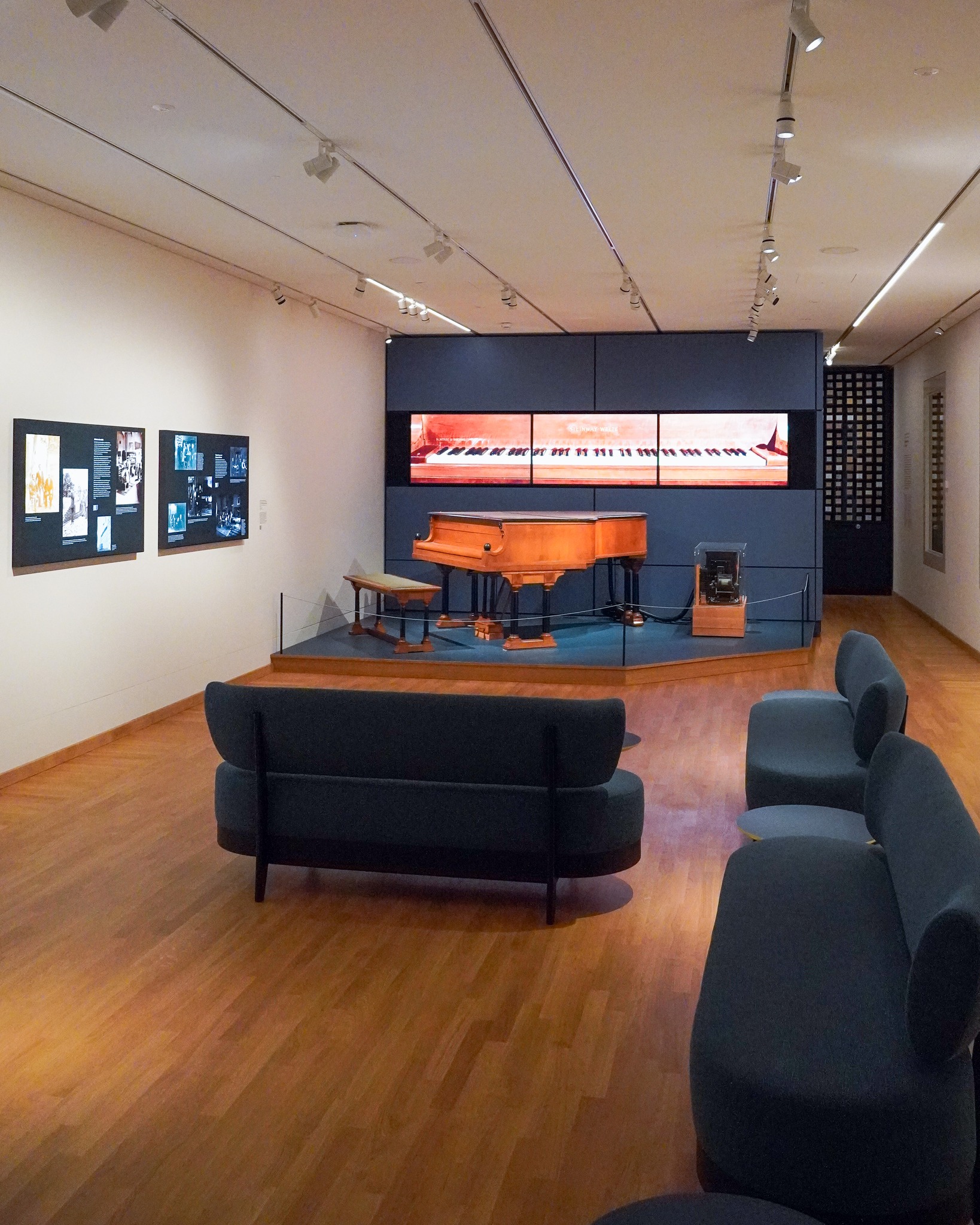 Ein Ausstellungsraum mit Holzboden und dunklen Sitzmöbeln im Vordergrund. Auf einer erhöhten Plattform steht ein historisches Klavier mit Hocker, flankiert von einem kleinen Exponat in einer Vitrine. Die Rückwand ist blau und zeigt einen langen Bildschirm mit Klaviertasten, während an der linken Wand Informationsplakate hängen. (KI-Alternativtext)