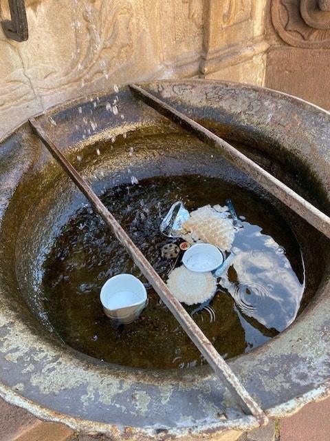 Ein alter, runder Brunnen mit Wasser, in dem mehrere leere Plastikbecher und ein Schaumstoffstück treiben. Zwei Metallstäbe liegen diagonal über dem Becken, im Hintergrund ist eine verzierte Steinwand zu sehen. (KI-Alternativtext)