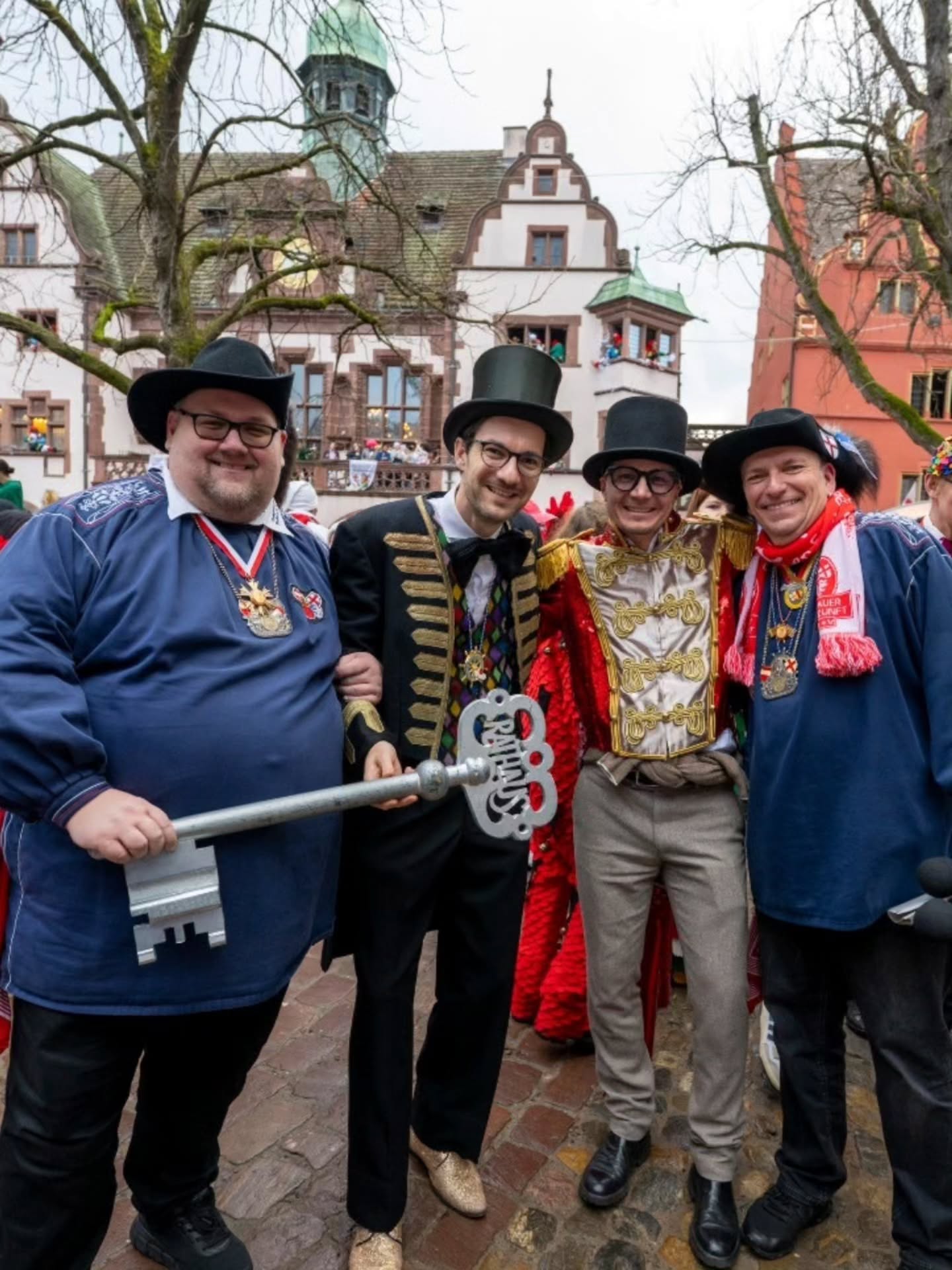 Dieser Alternativtext wurde KI-generiert: Mehrere Personen mit auffälligen Kostümen und Hüten stehen vor einem Gebäude mit Fachwerkelementen. Im Hintergrund sind weitere Menschen und festliche Dekorationen zu sehen.